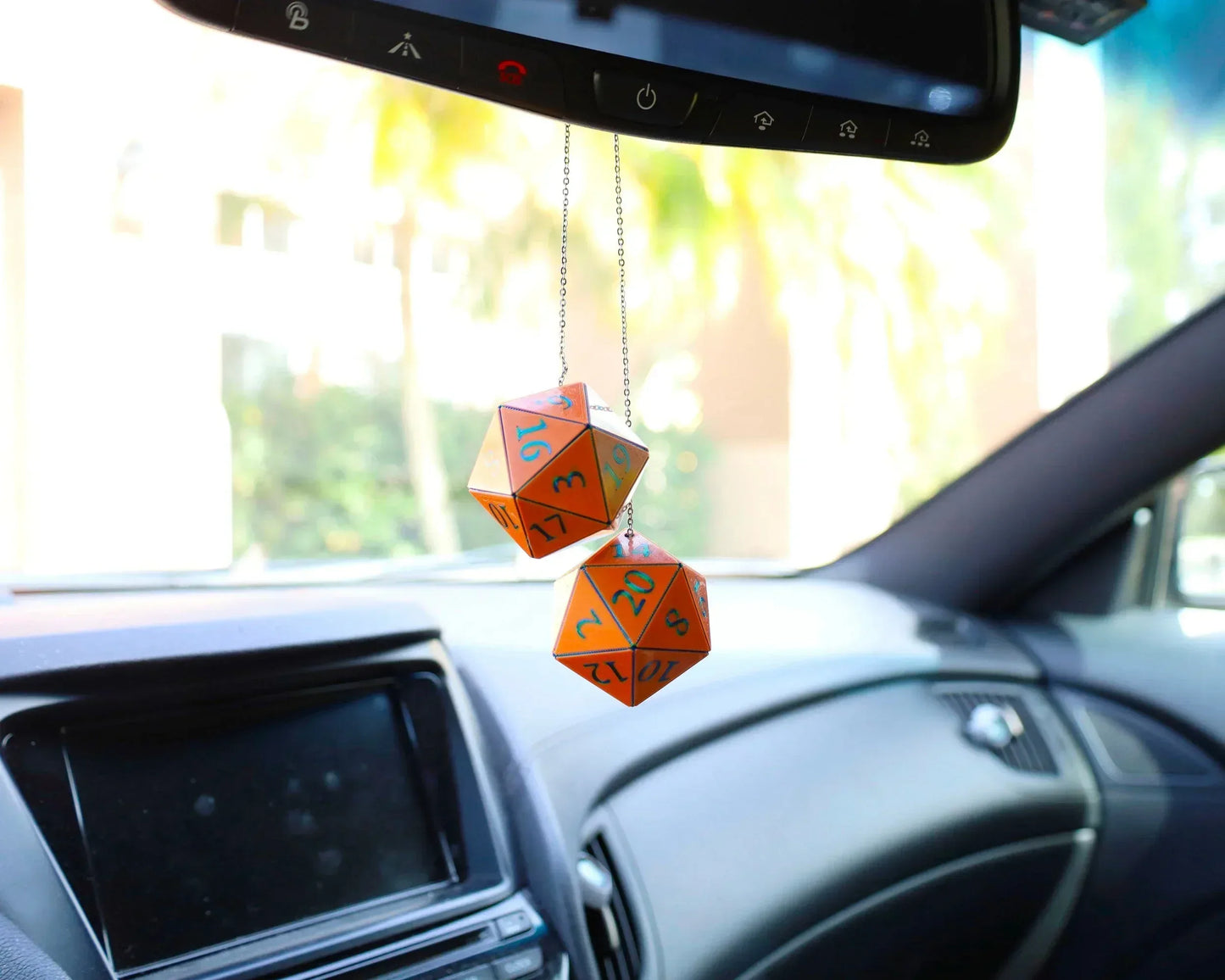 Orange twenty-sided dice car charm hanging from rearview mirror in a sunlit vehicle interior.