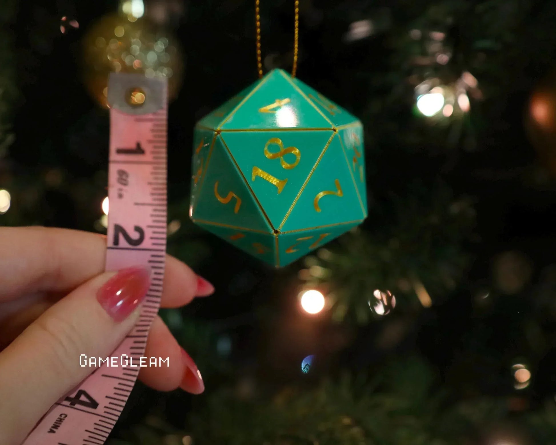 Green 20-sided dice ornament with gold numbers hanging on Christmas tree, measured for size.