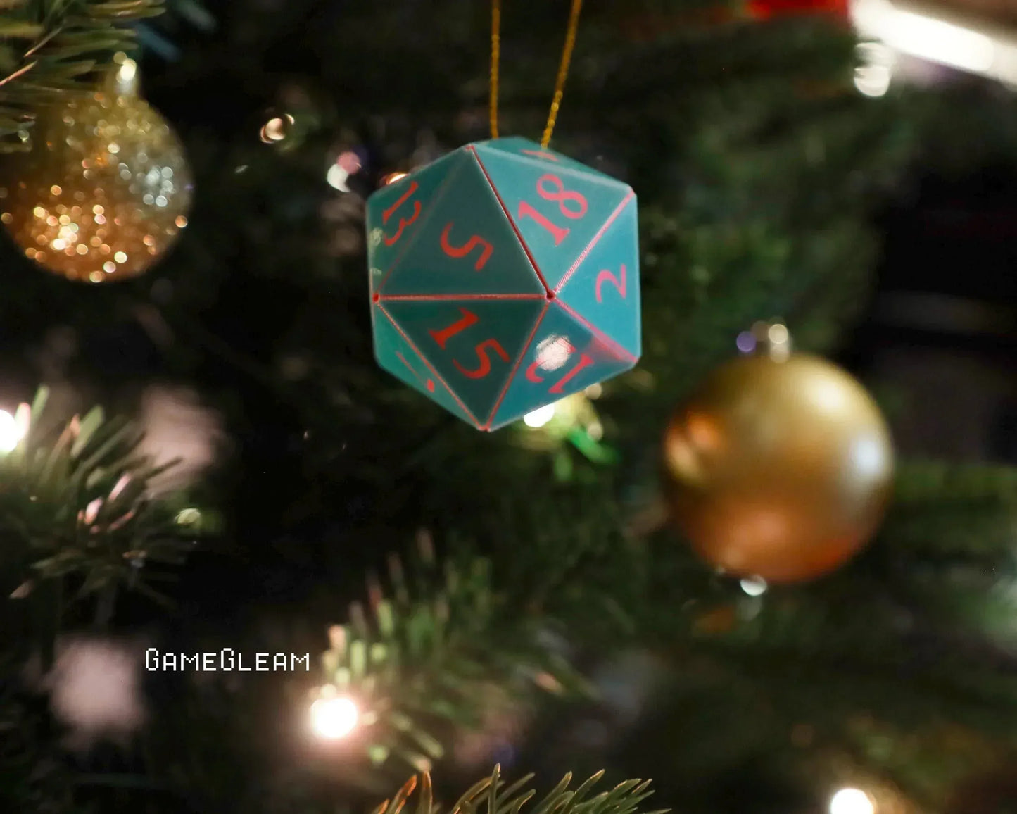 Blue twenty-sided dice ornament with red numbers hanging on a Christmas tree with gold baubles.