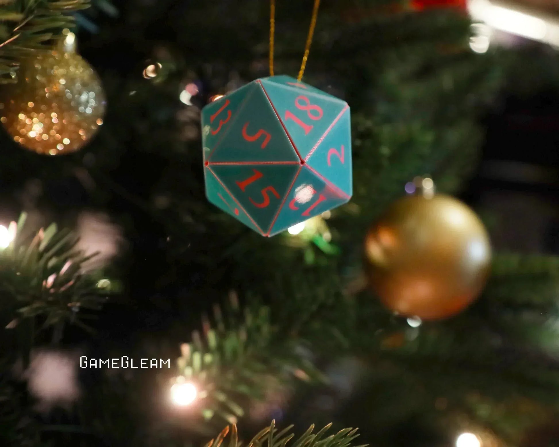 Blue twenty-sided dice ornament with red numbers hanging on a Christmas tree with gold baubles.