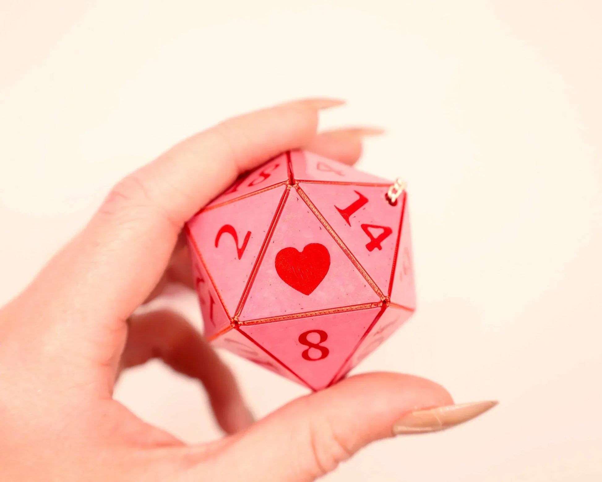Hand holding a pink 20-sided dice charm with red numbers and heart symbol, gamer accessory