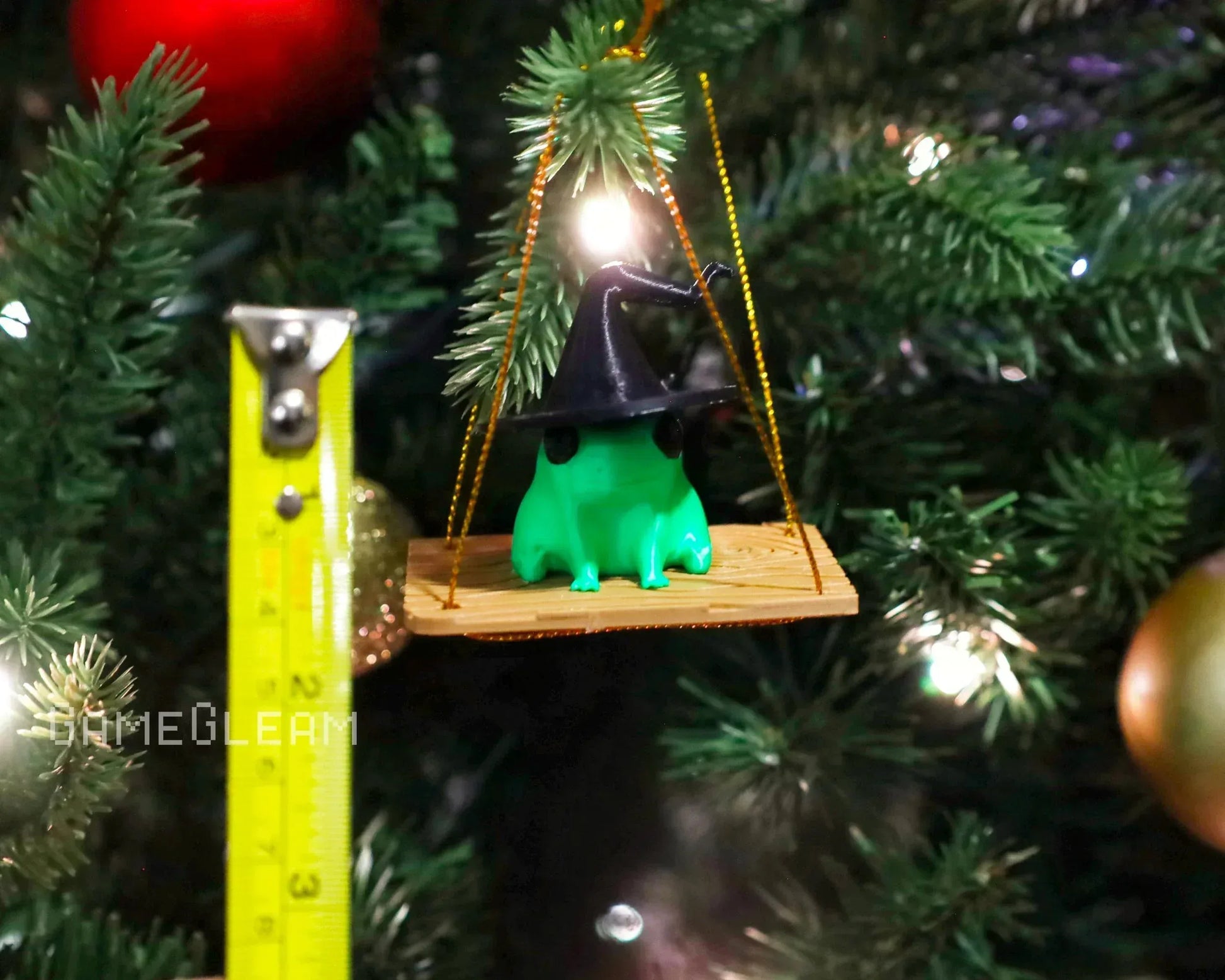 Green frog ornament with a witch hat on a swing, hanging on a Christmas tree branch.