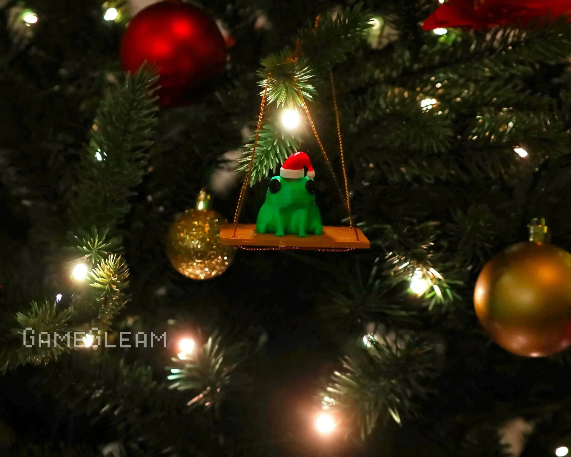 Green frog ornament with Santa hat sitting on a swing, hanging on a decorated Christmas tree.