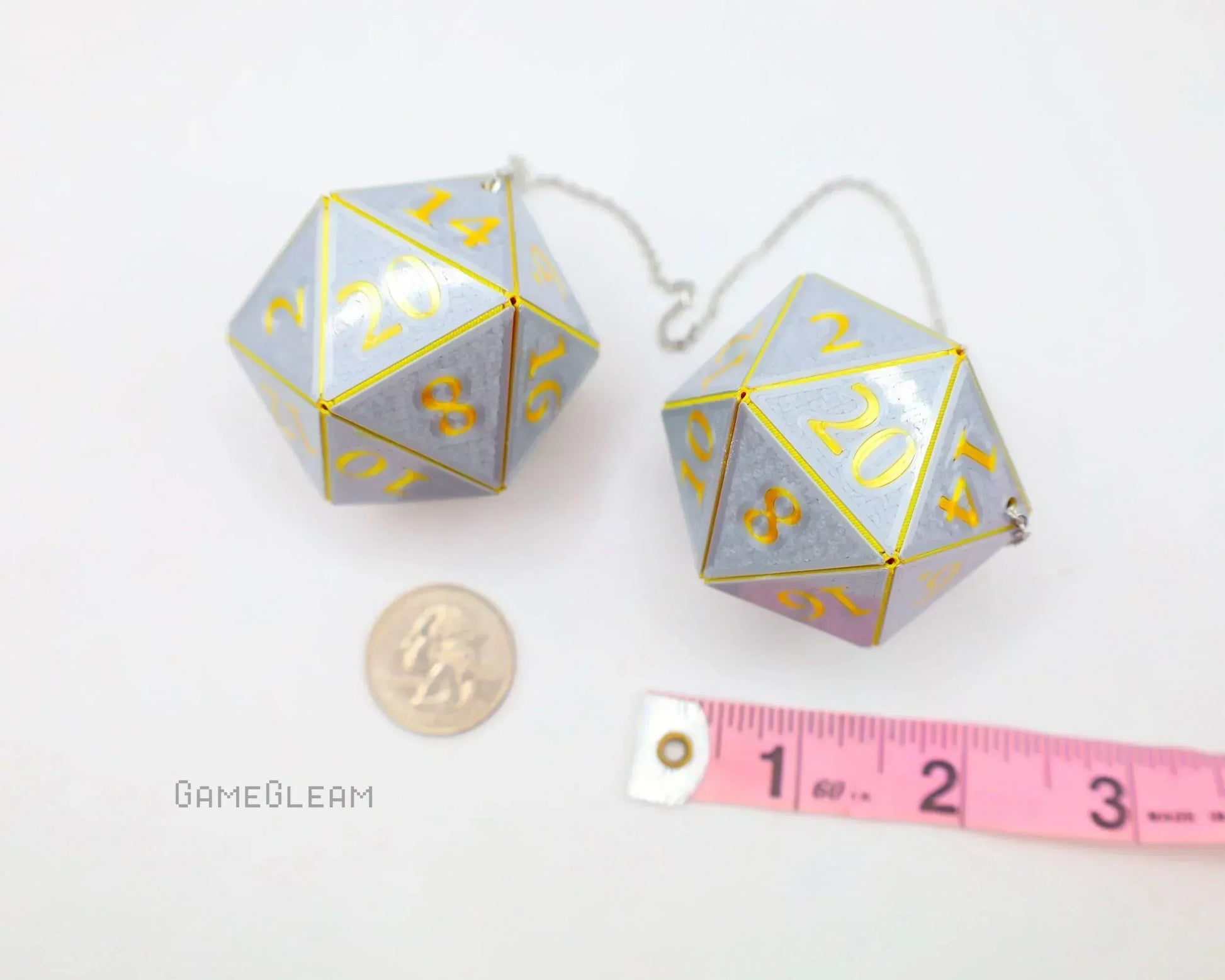 Pair of large silver 20-sided dice charms with yellow numbers, next to a ruler and coin