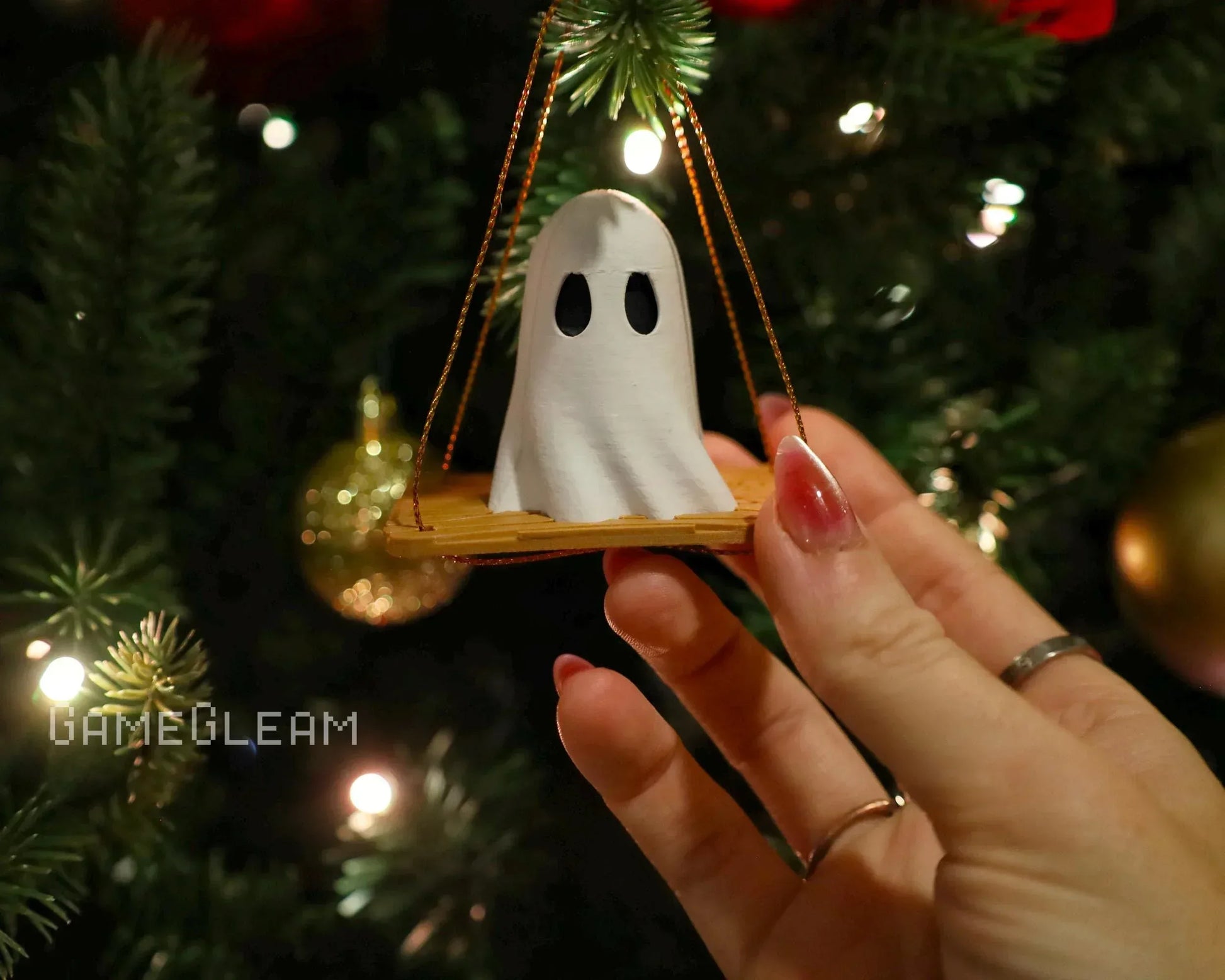 Hand holding cute ghost ornament on swing, hanging on Christmas tree with lights and gold baubles