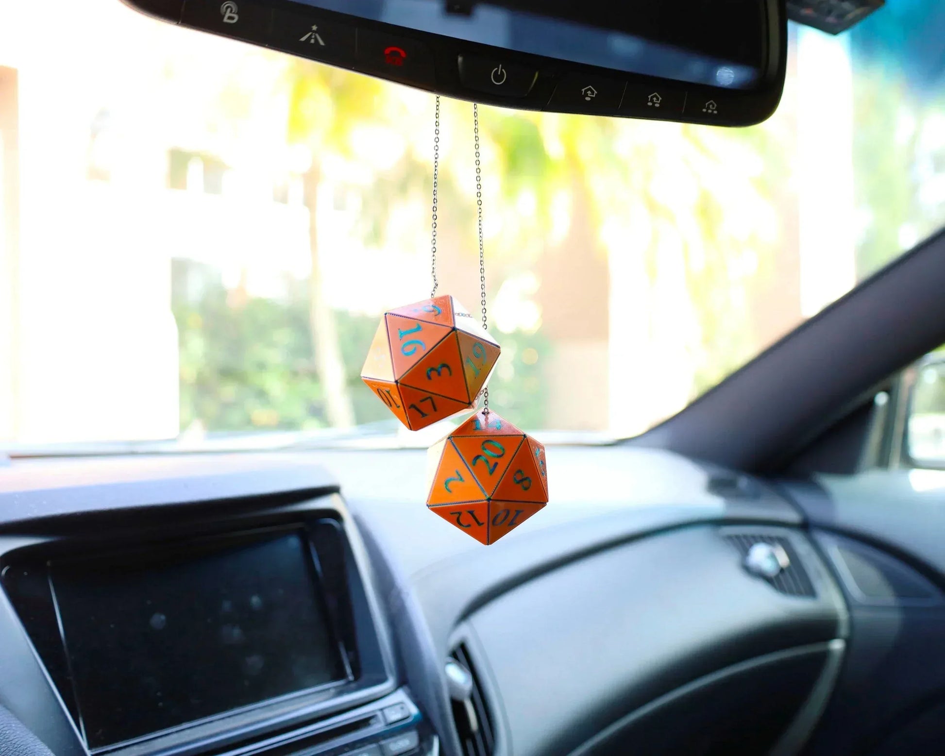 Orange twenty-sided dice car charm hanging from rearview mirror in a sunlit vehicle interior.