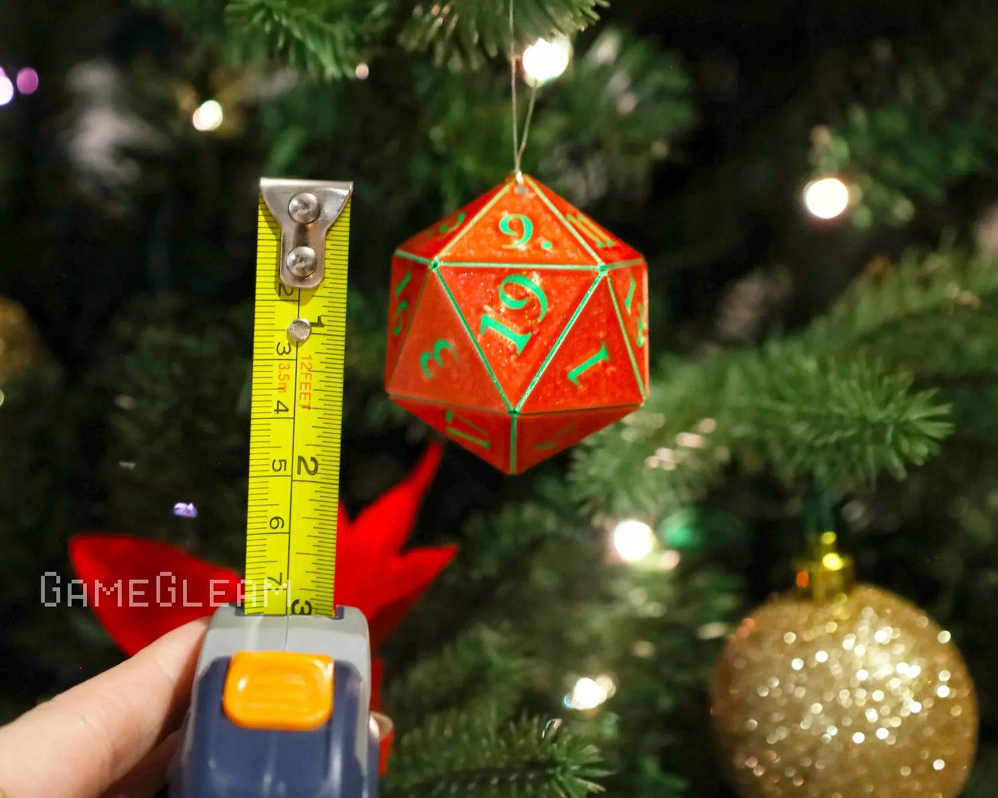Red 20-sided dice ornament with green numbers hanging on a Christmas tree, tape measure beside it.