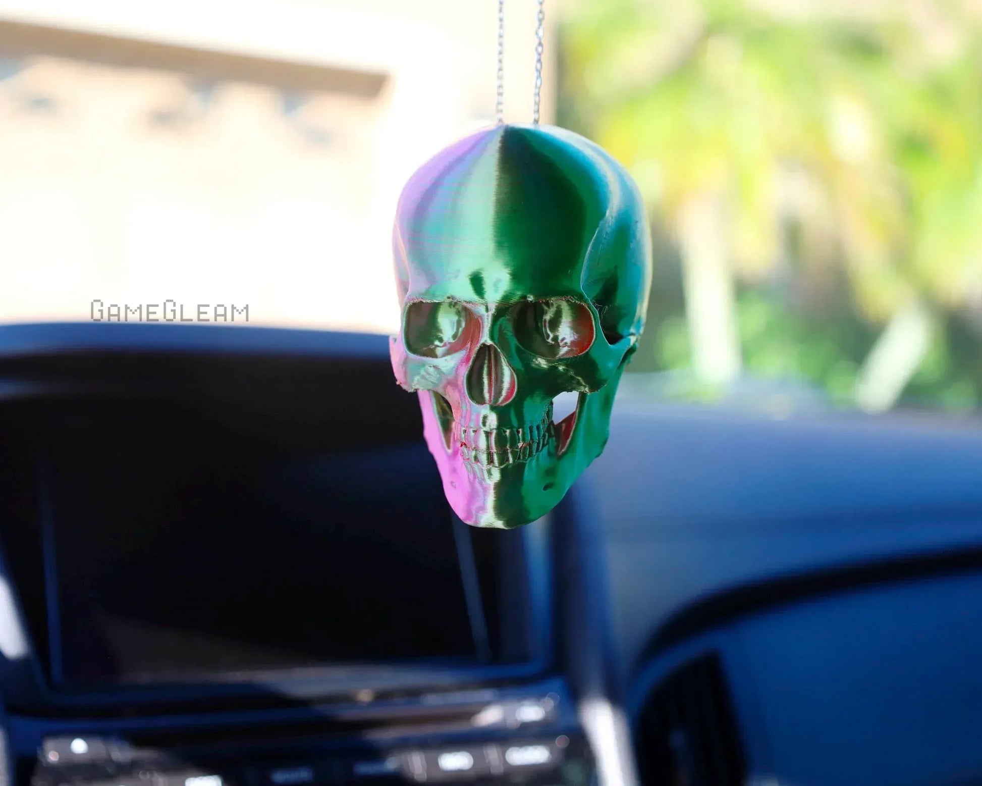 Holographic green and pink skull car charm hanging from rearview mirror in sunlight