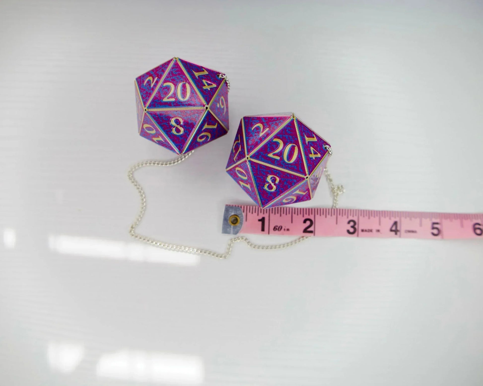 Holographic purple 20-sided dice charms with gold numbers and chain, next to pink measuring tape