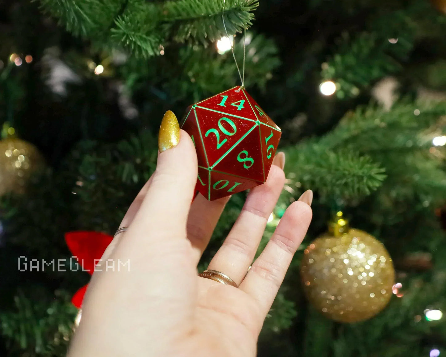 Hand holding red twenty-sided dice ornament with green numbers near Christmas tree