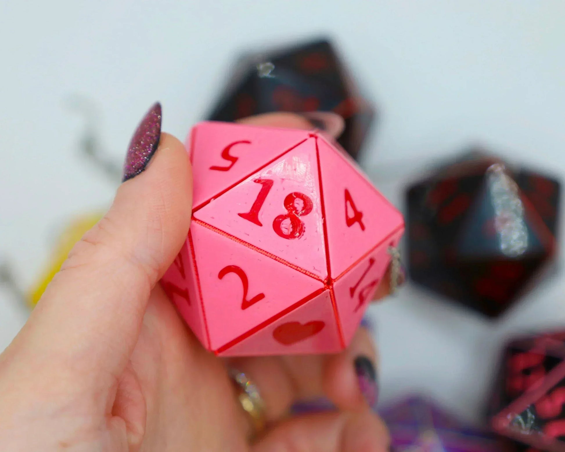 Hand holding a pink handmade 20-sided dice charm with red numbers for tabletop gaming.