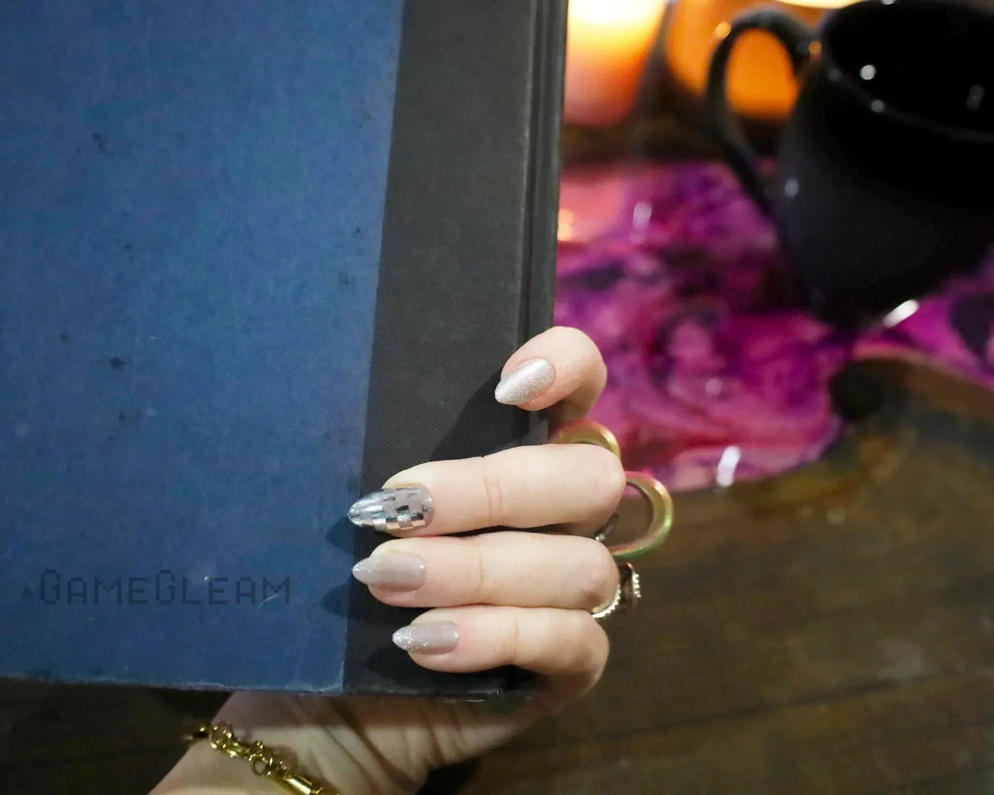 Hand with metallic gamer-themed nails holding a blue book, candles and mug in background