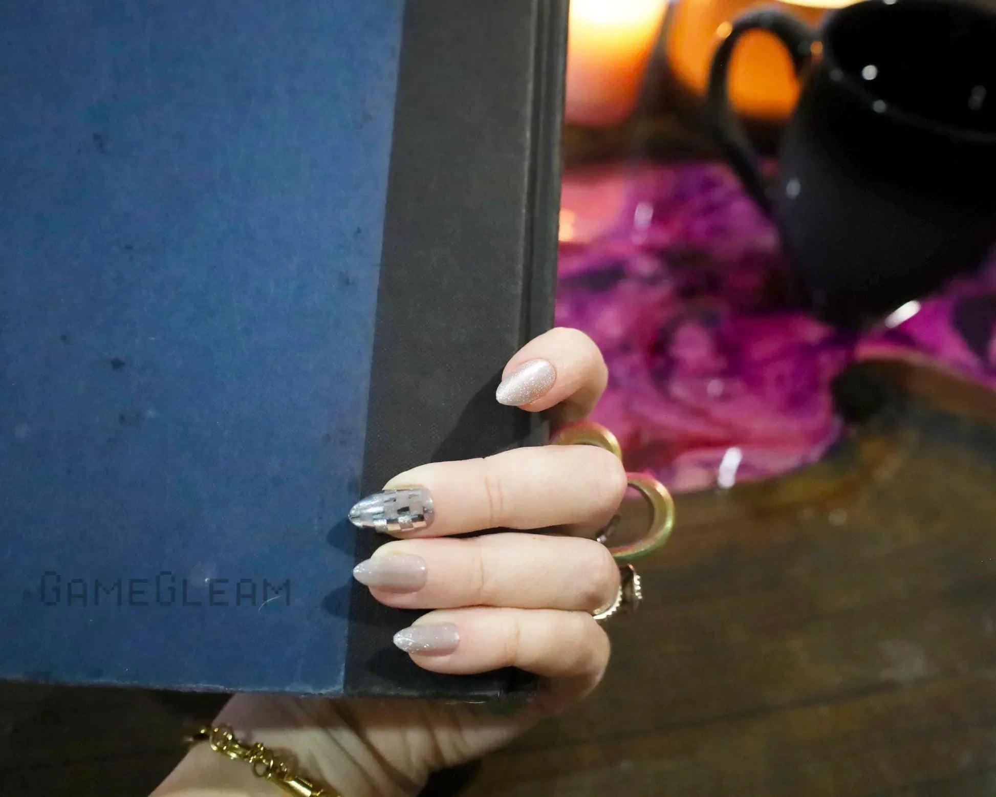 Hand with metallic gamer-themed nails holding a blue book, candles and mug in background