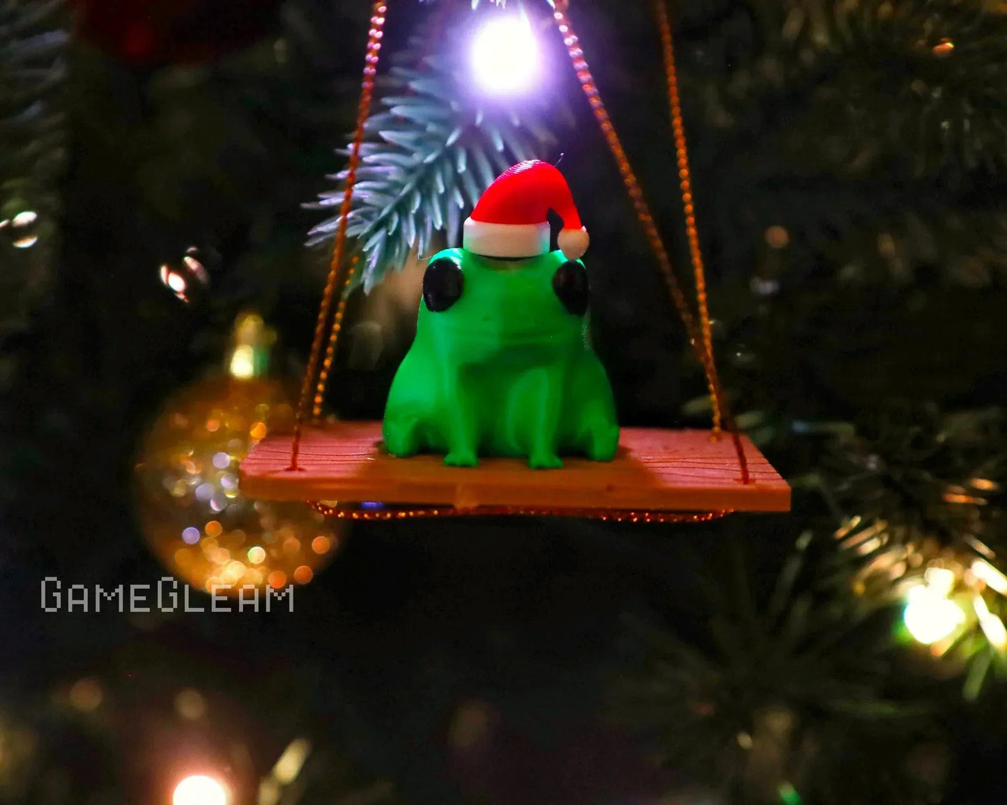 Green frog ornament with Santa hat sitting on a swing, hanging from a Christmas tree.