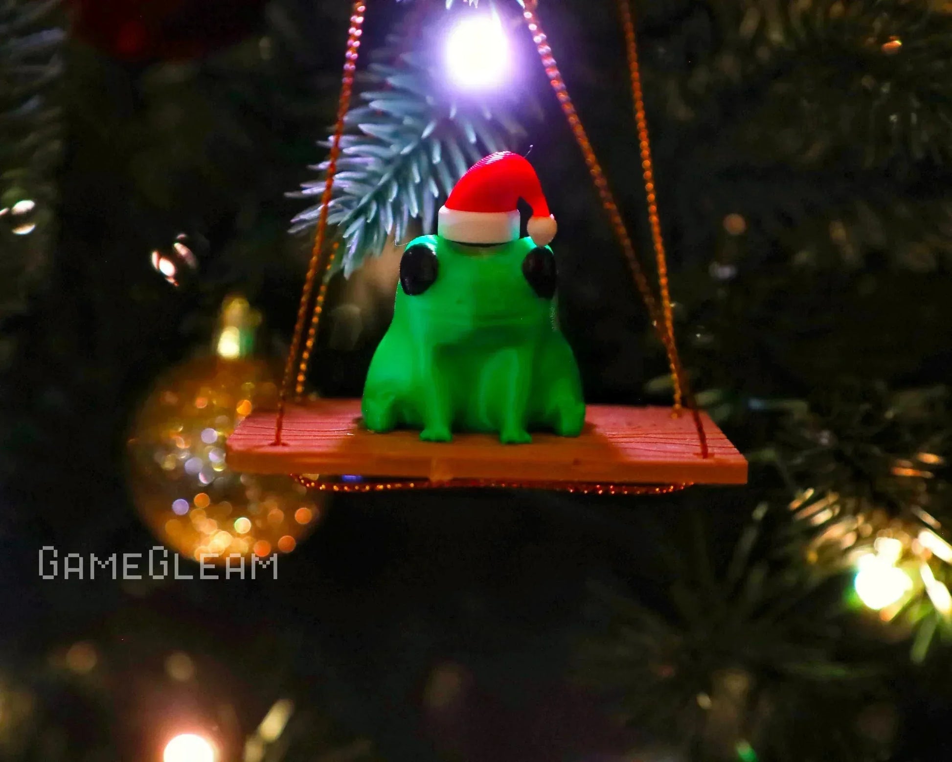 Green frog ornament with Santa hat sitting on a swing, hanging from a Christmas tree.