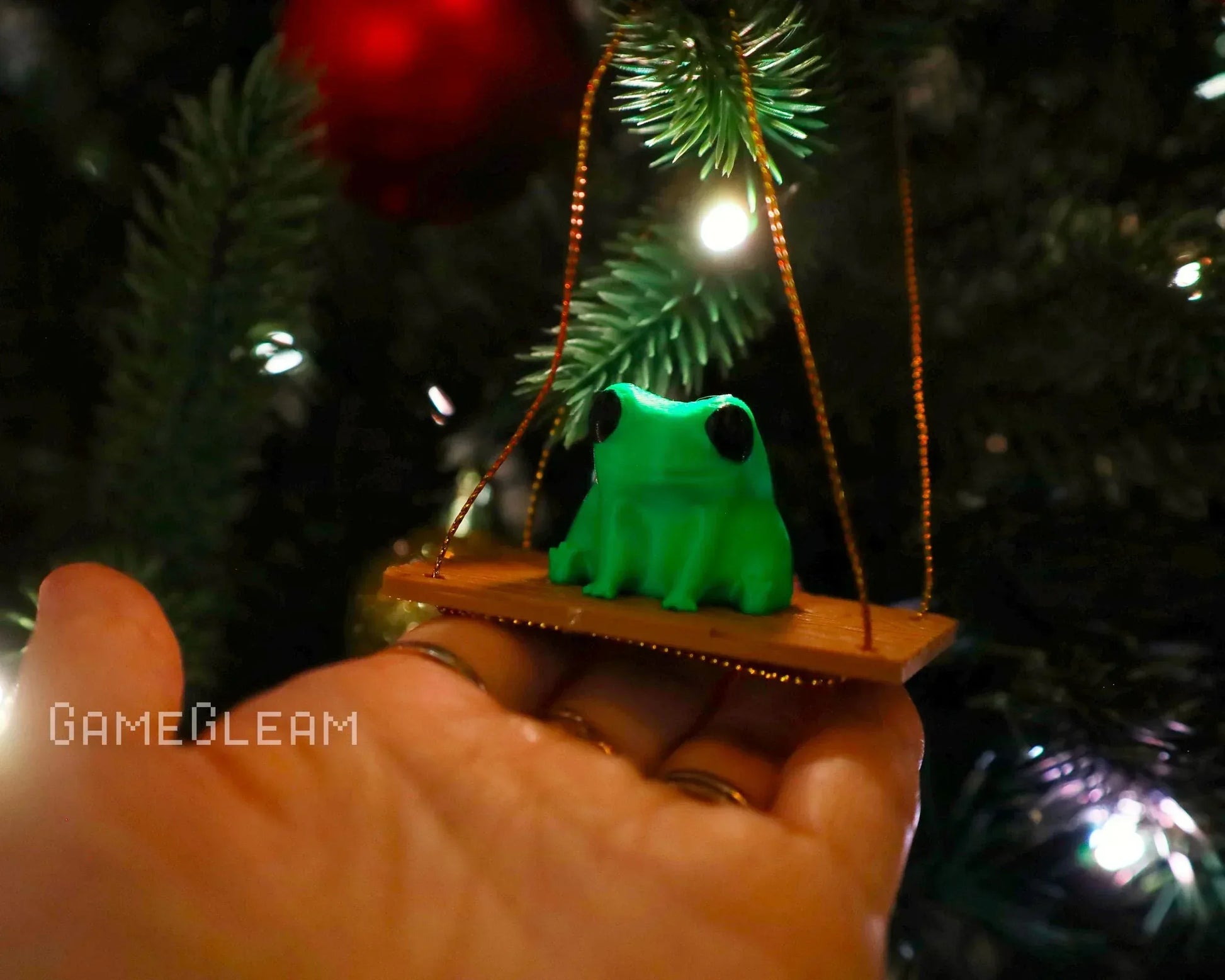 Hand holding a green frog ornament on a swing, hanging from a Christmas tree branch.