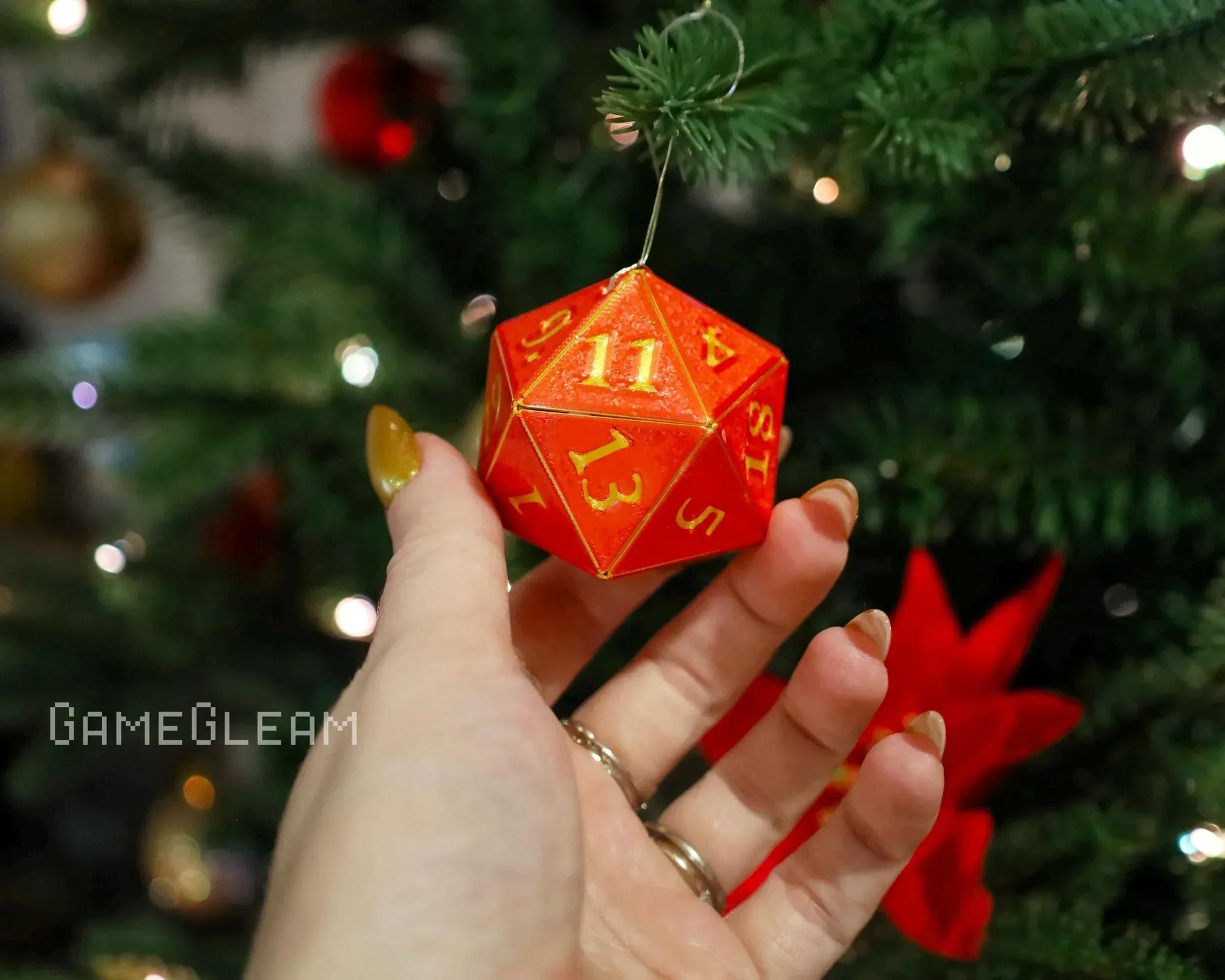 Hand holding a red glittery twenty-sided dice ornament with yellow numbers near a Christmas tree.