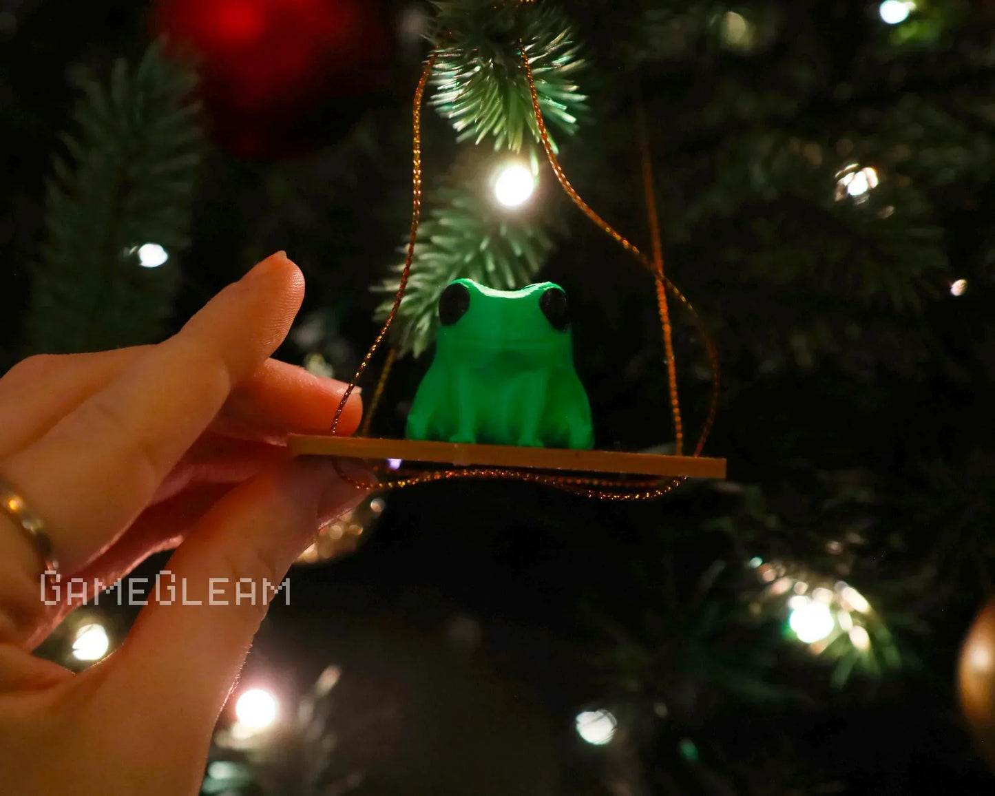 Hand holding a green frog ornament on a swing, hanging on a lit Christmas tree branch.
