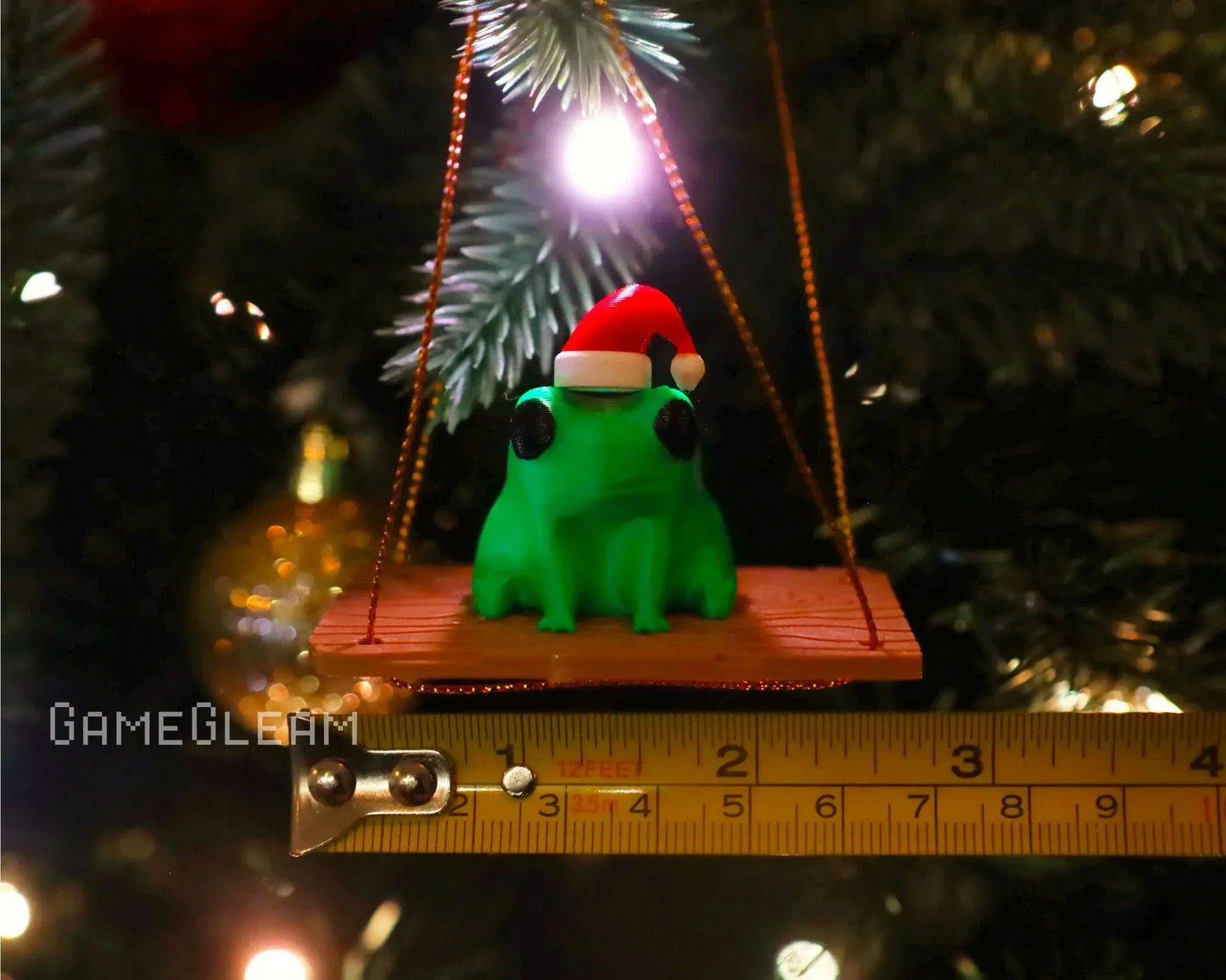 Green frog ornament with Santa hat sitting on a swing, hanging on a Christmas tree branch.