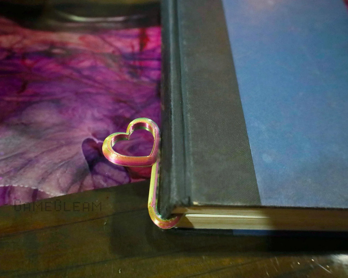 Iridescent heart-shaped metal bookmark clipped to a book on a purple marbled surface