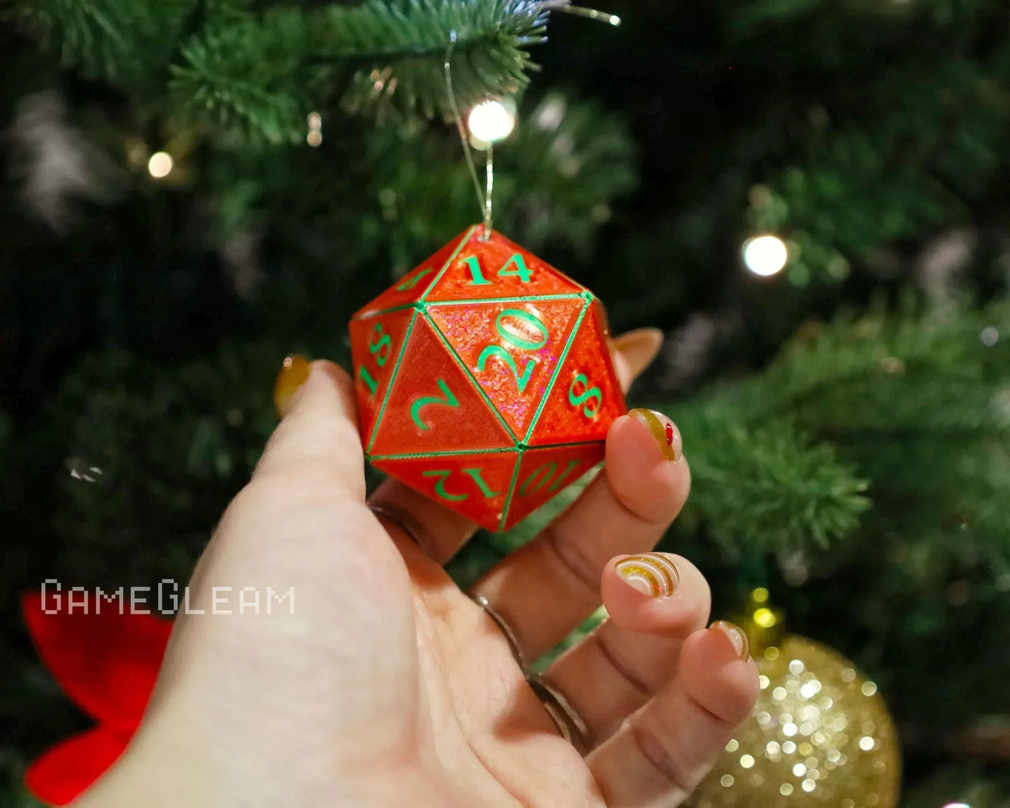 Hand holding a red glittery 20-sided dice ornament with green numbers near a Christmas tree.