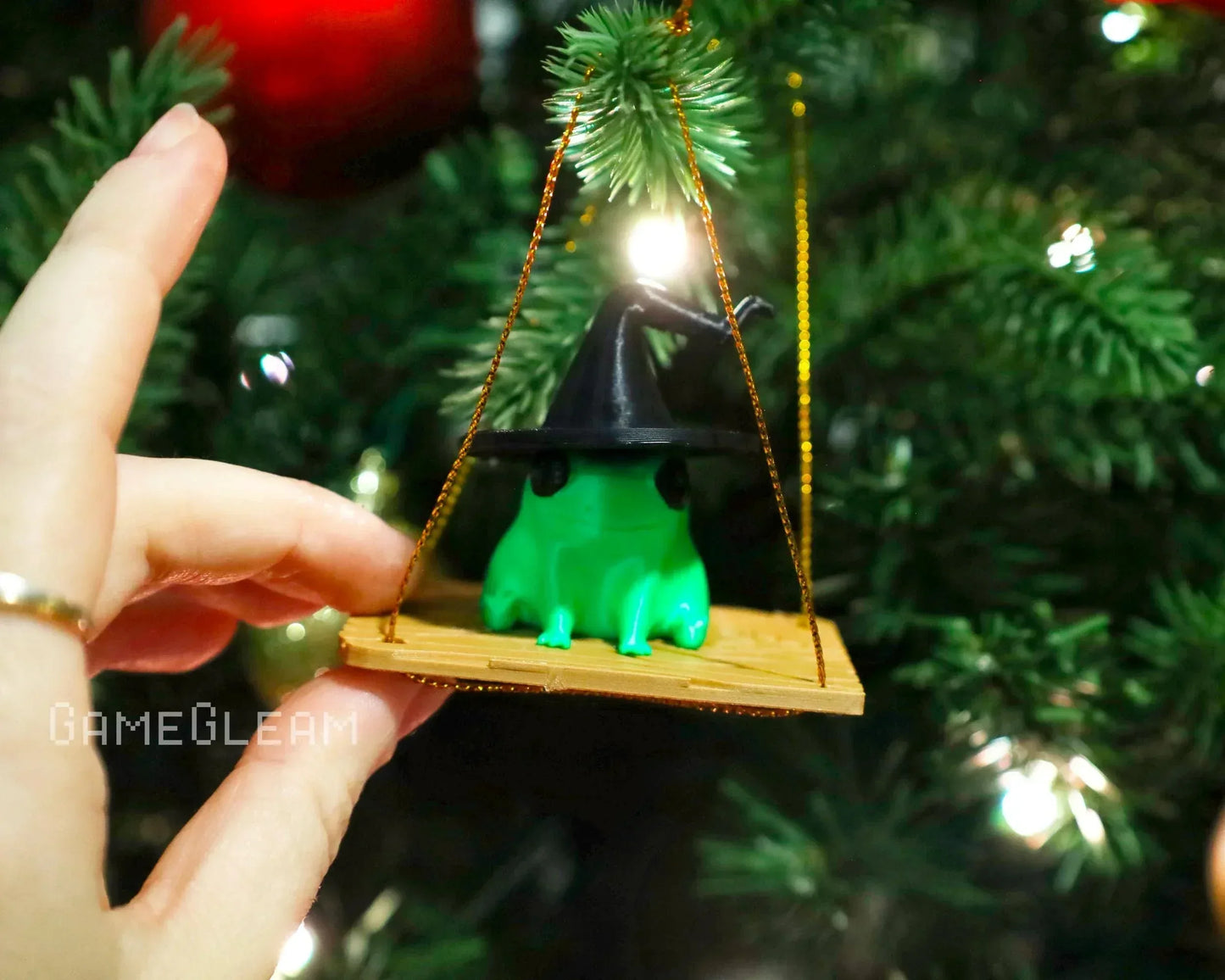 Hand holding green frog figurine with wizard hat on wooden swing, hanging on Christmas tree