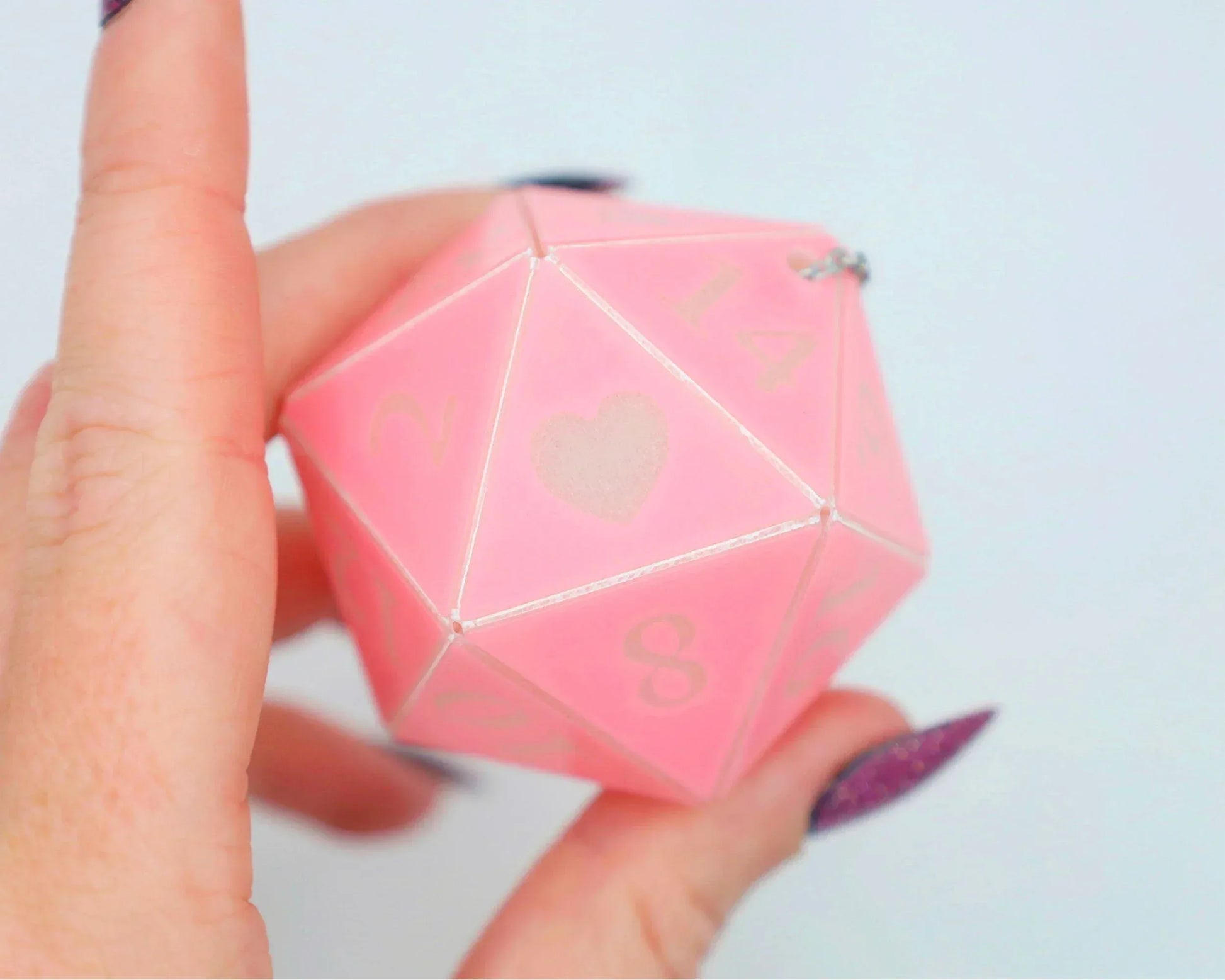 Hand holding a large pink 20-sided dice charm with heart and numbers, pastel aesthetic.