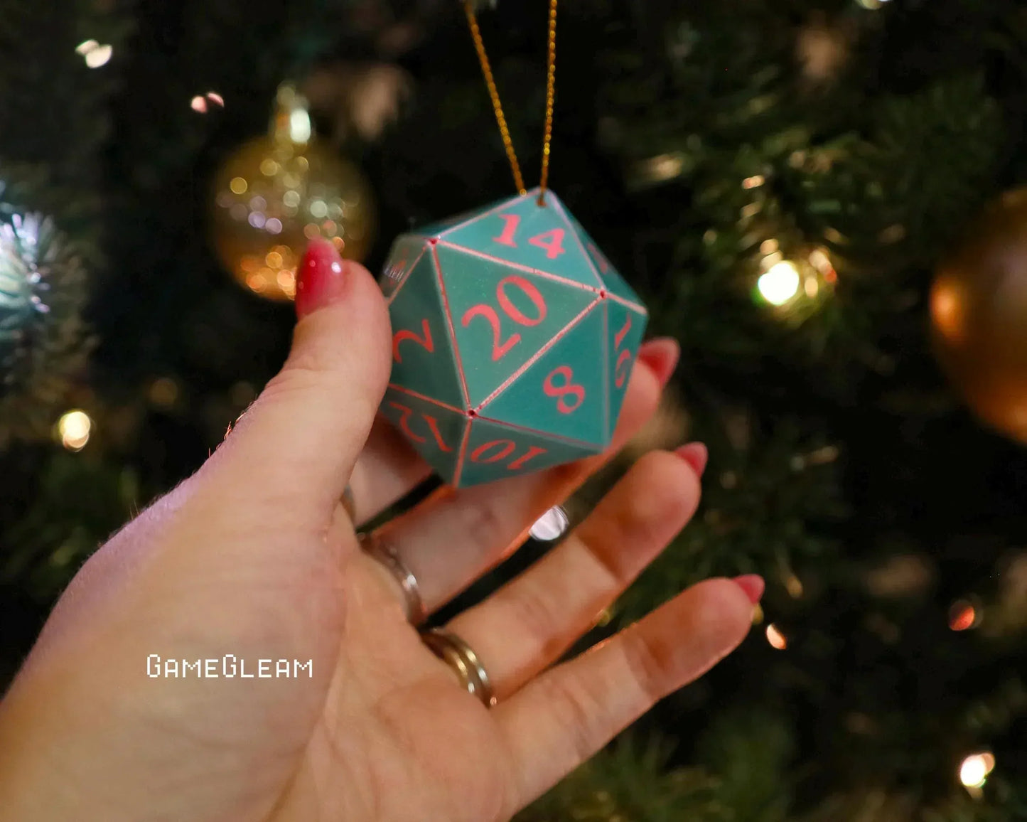 Hand holding teal 20-sided dice ornament with pink numbers near a decorated Christmas tree.