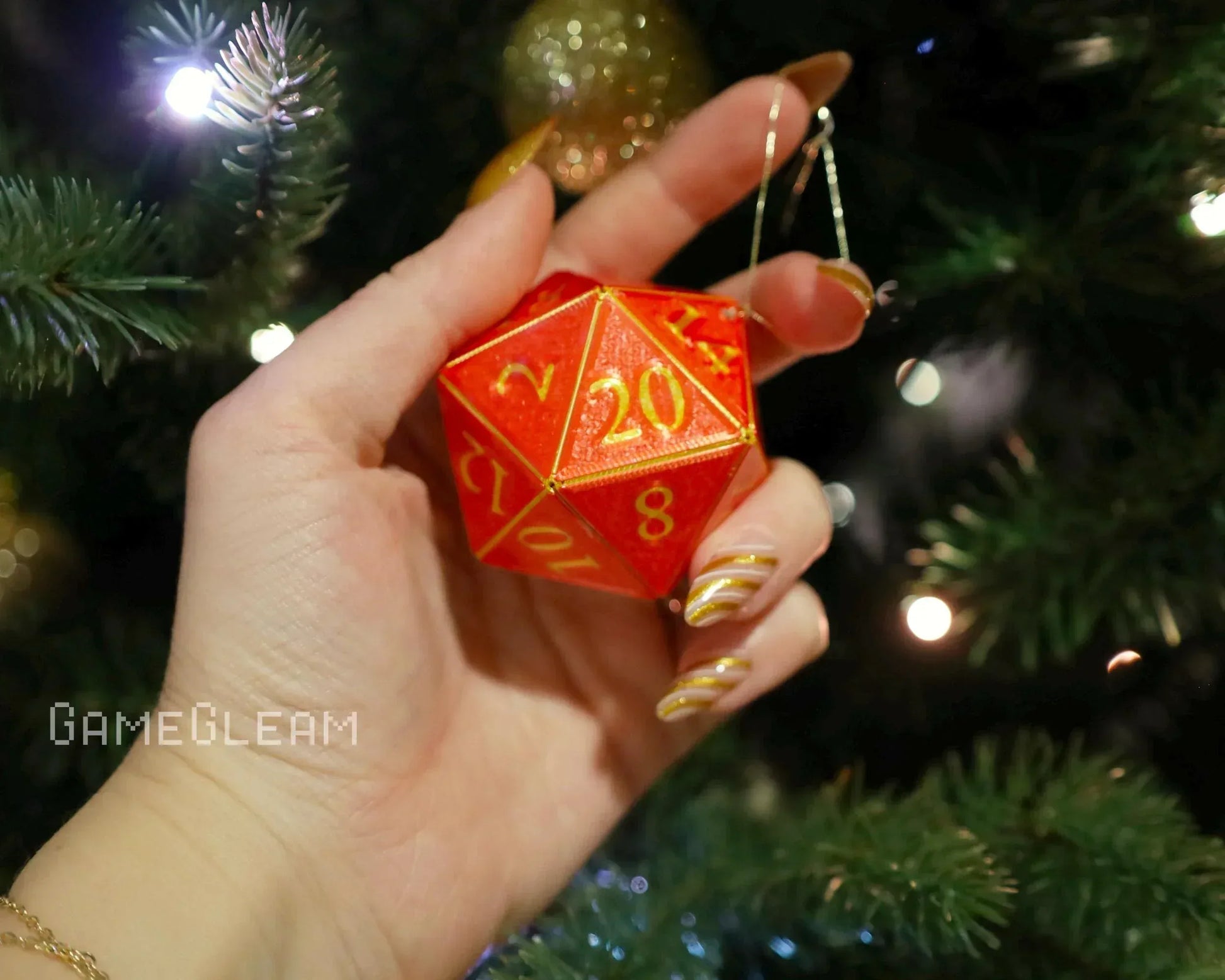 Hand holding a red twenty-sided dice ornament with gold numbers near a decorated Christmas tree.