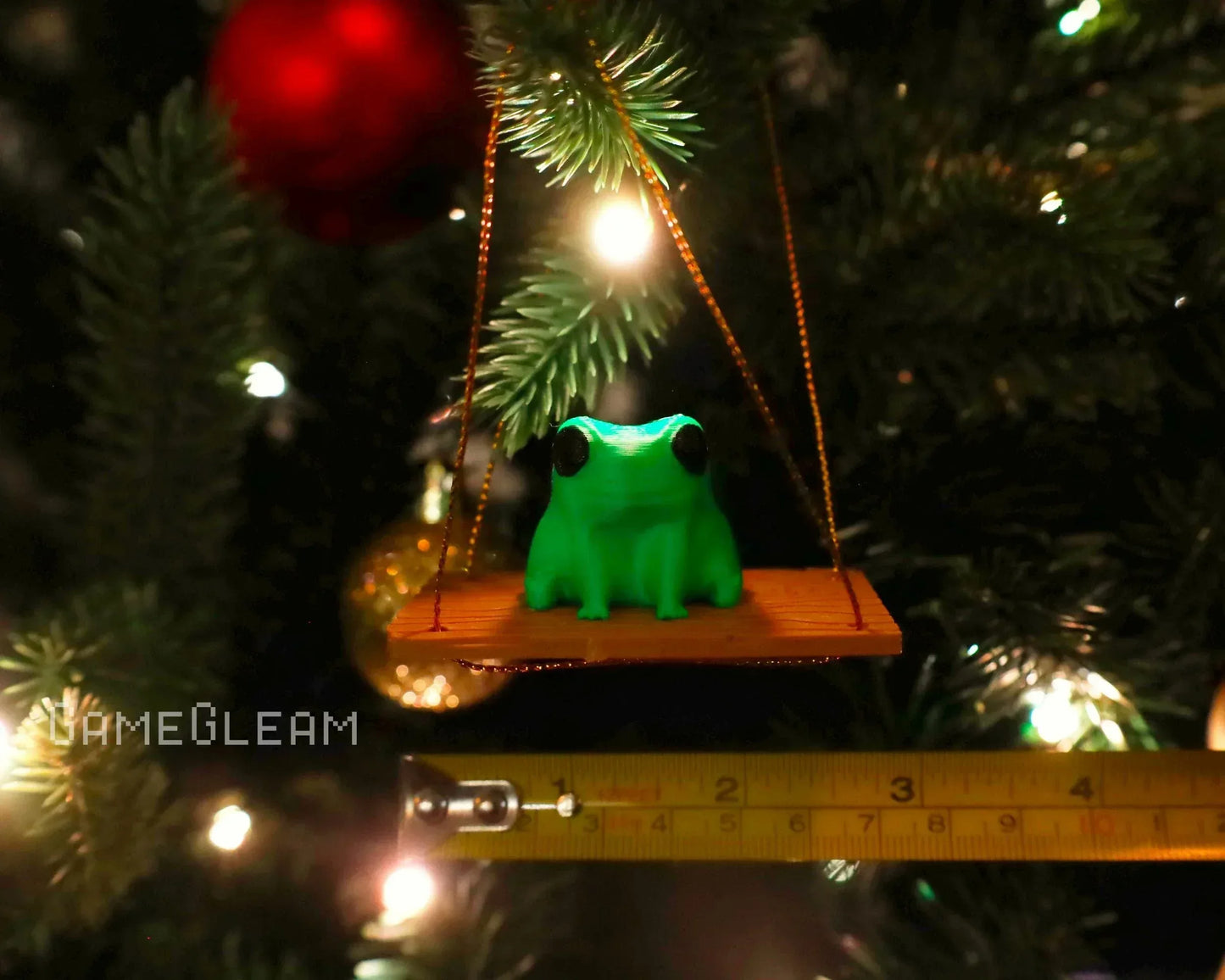 Green frog figurine on wooden swing ornament hanging from Christmas tree branch, with measuring tape