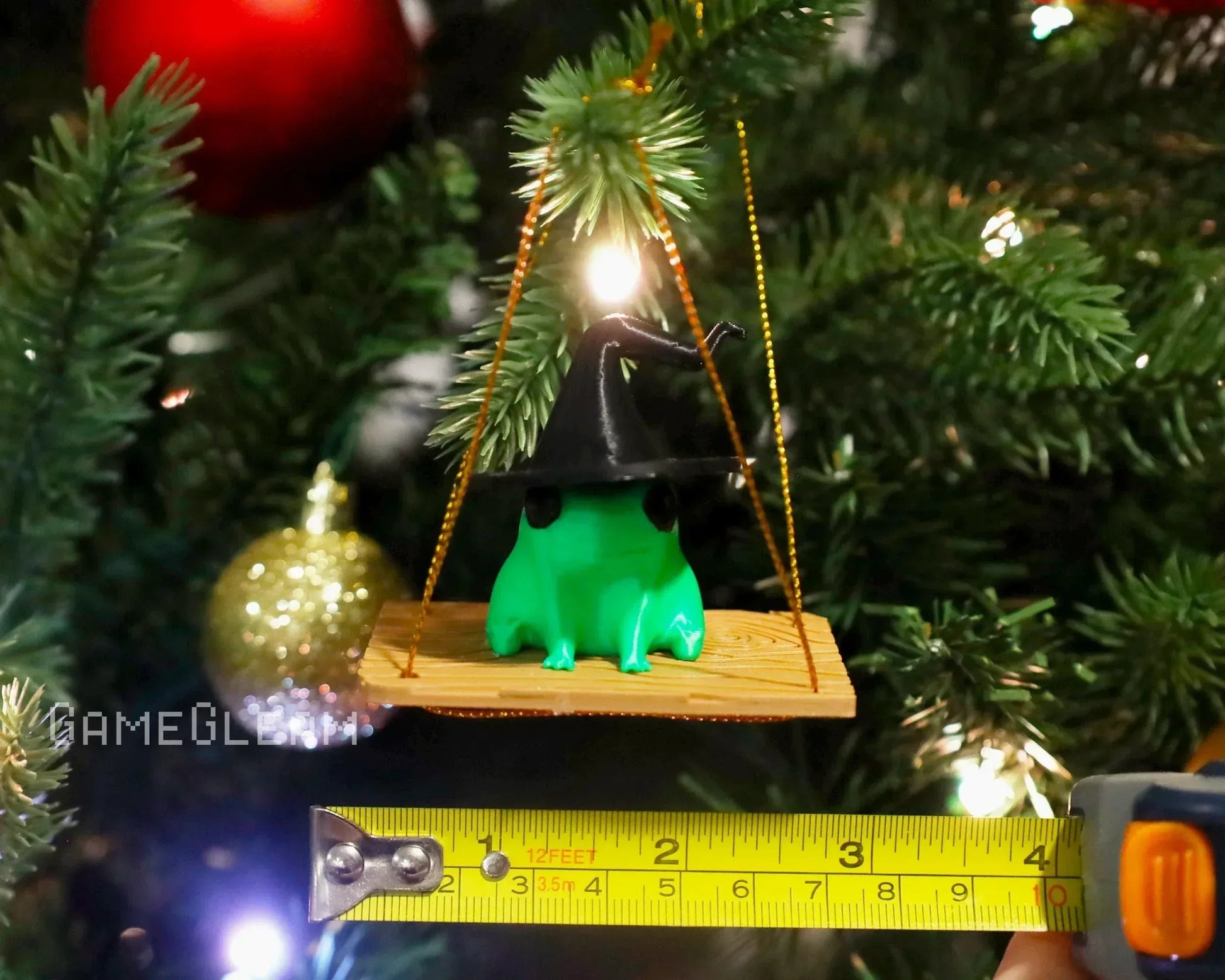 Green frog figurine with a black wizard hat sitting on a wooden swing Christmas ornament, measured with tape, hanging on a decorated tree.