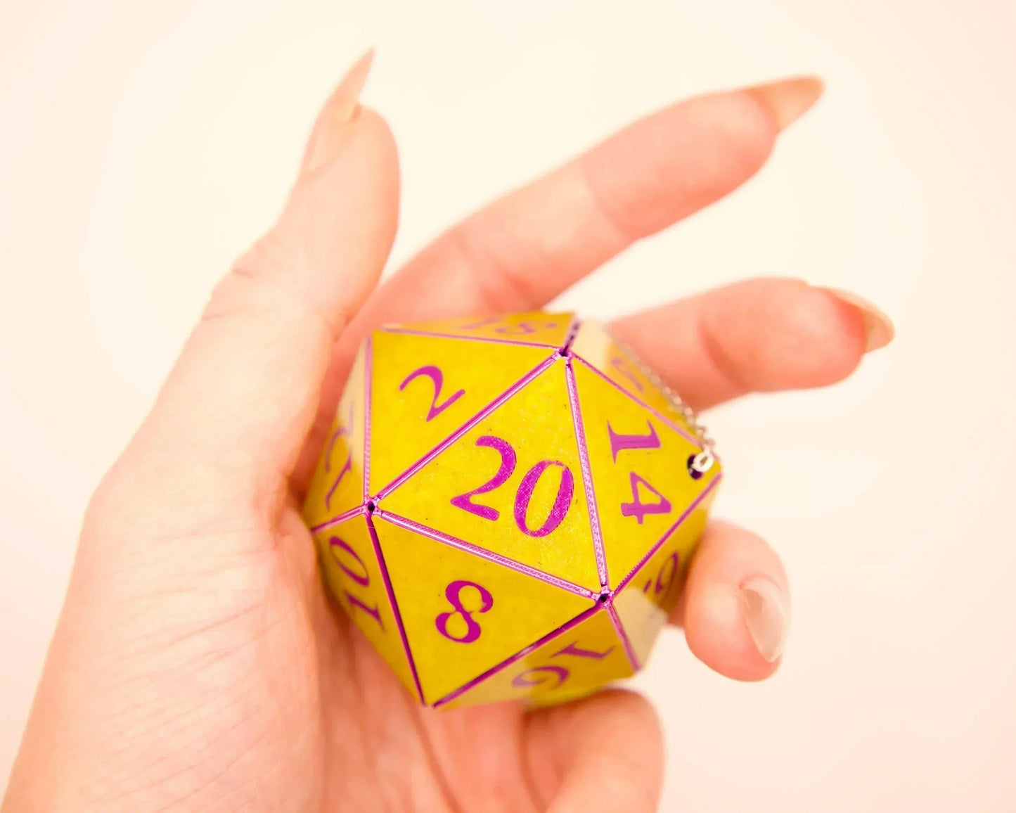 Hand holding a yellow twenty-sided dice charm with purple numbers, close-up view.
