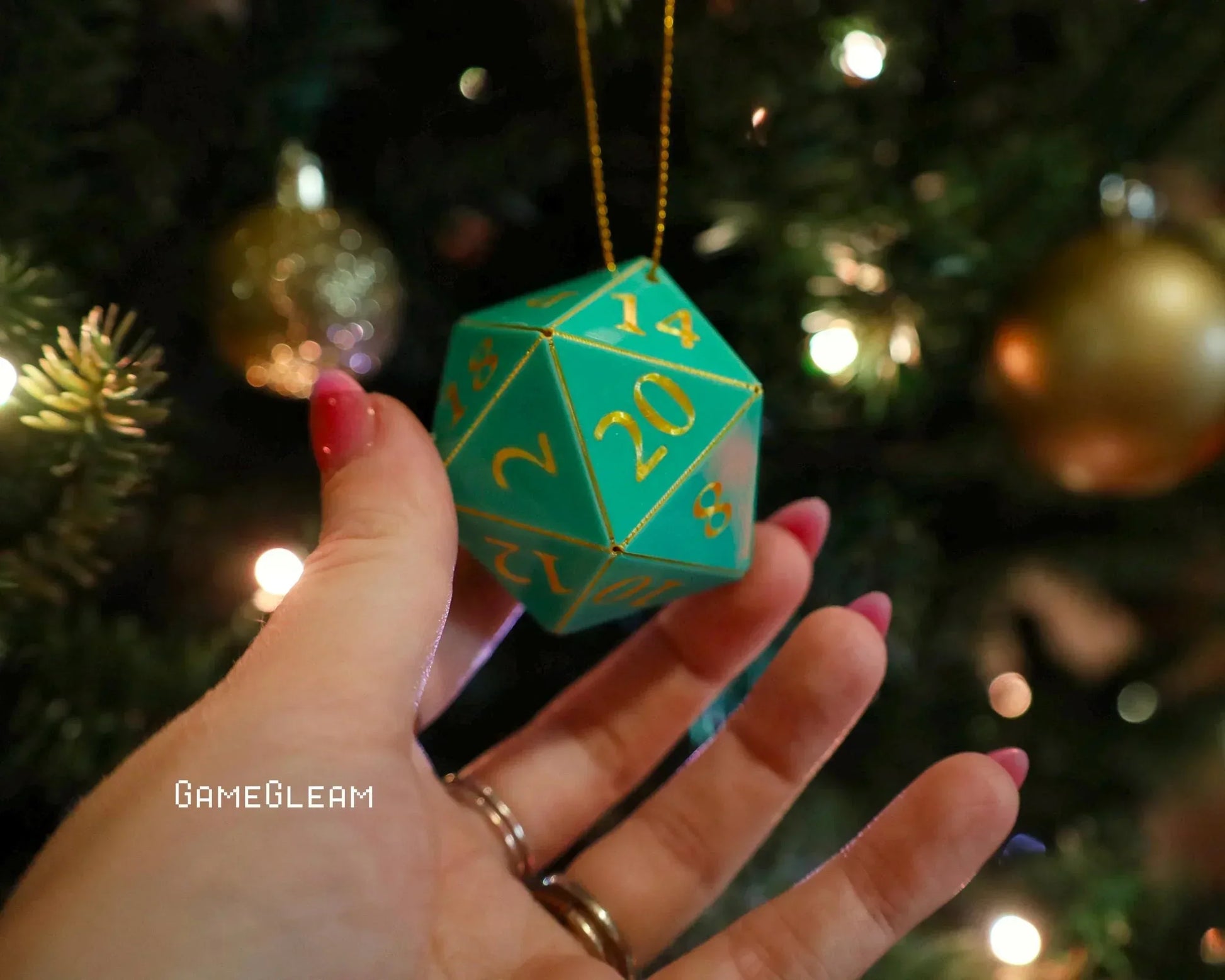 Hand holding green 20-sided dice ornament with gold numbers near a decorated Christmas tree.