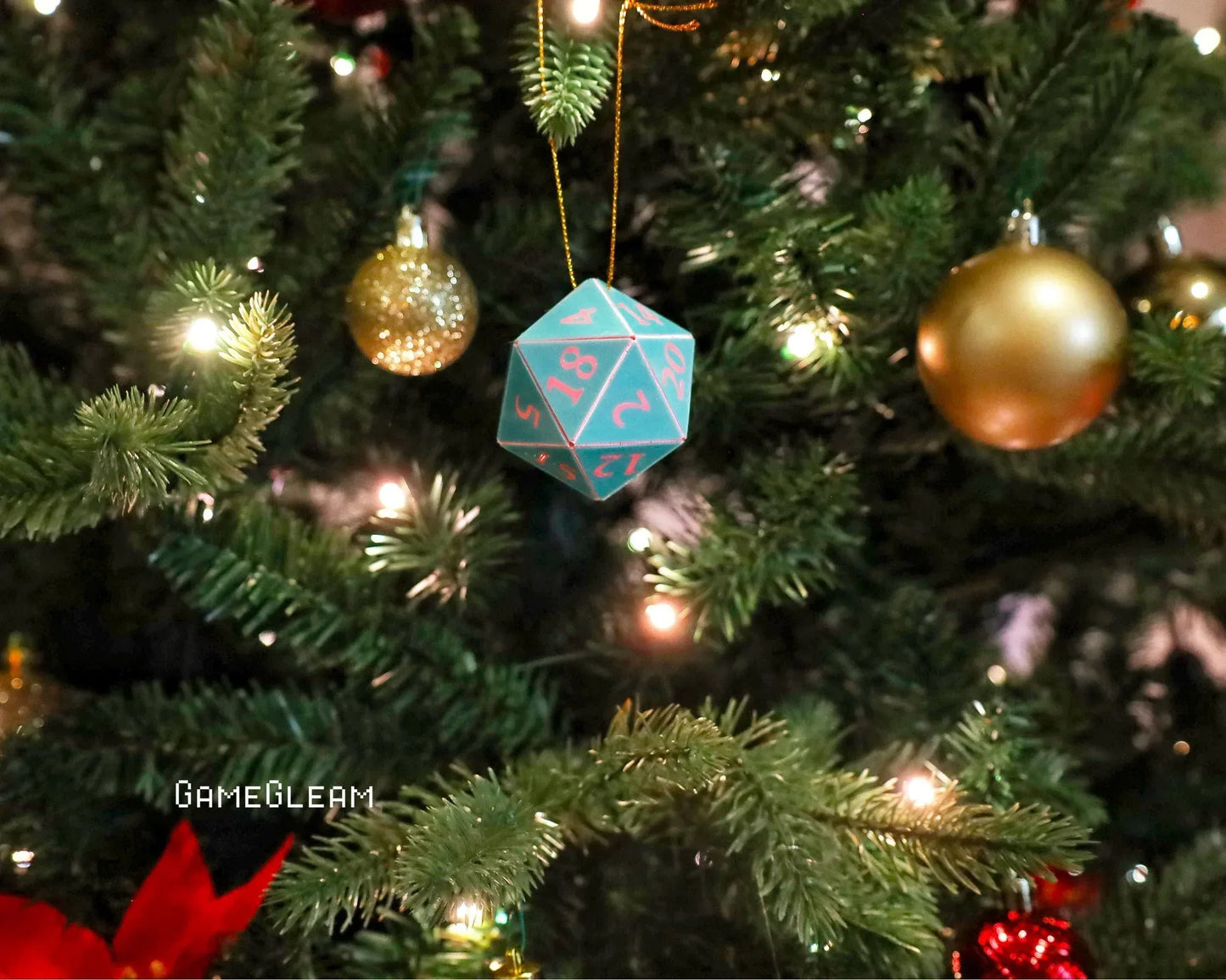 Turquoise and pink twenty-sided dice ornament hanging on a lit Christmas tree with gold baubles.
