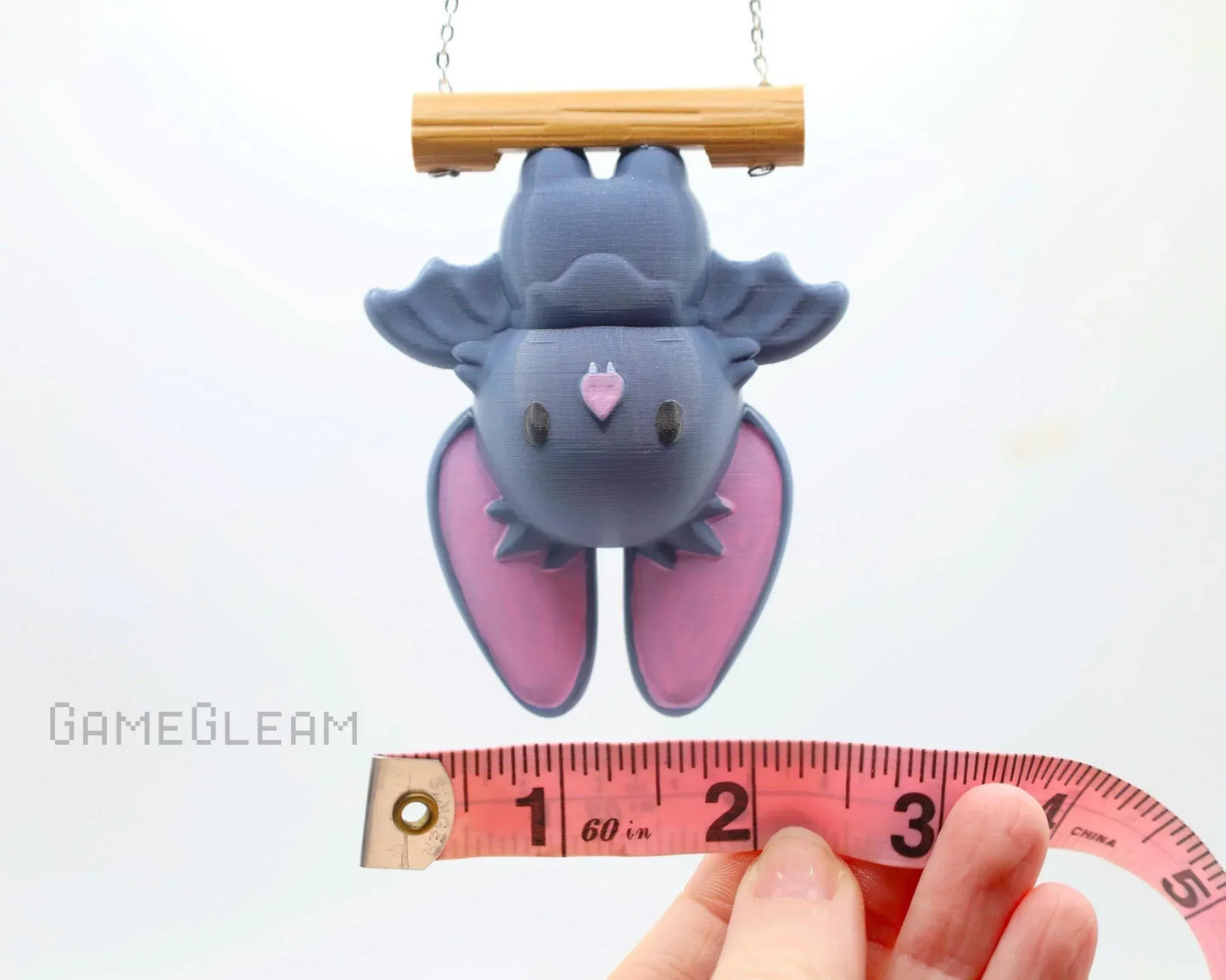 Cute handmade bat charm with purple wings hanging from a wooden perch, shown with measuring tape