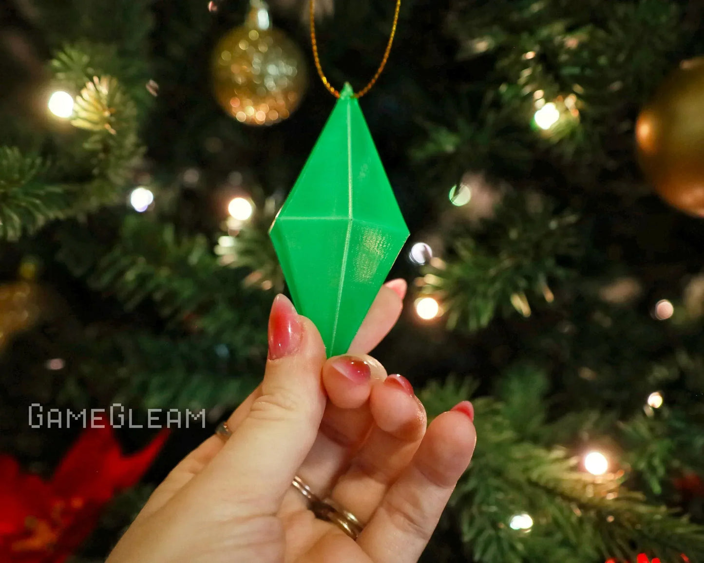 Hand holding green geometric ornament in front of a Christmas tree with gold baubles.