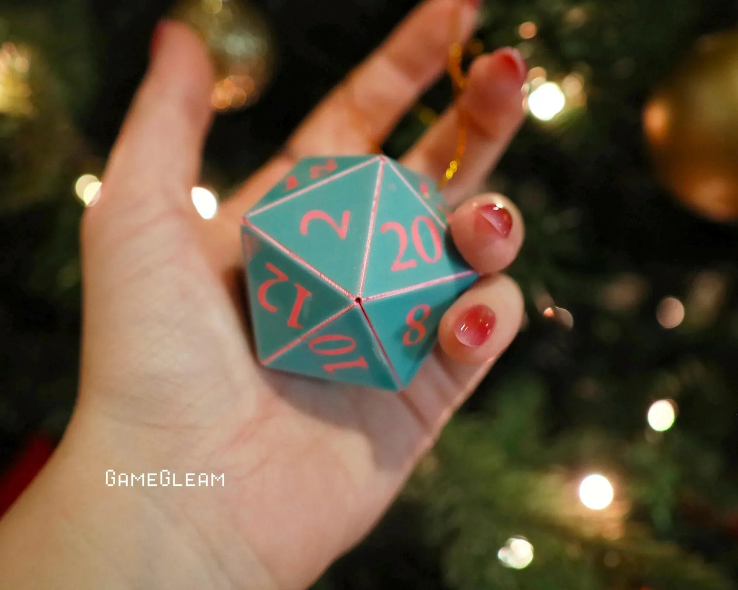 Hand holding teal handmade twenty-sided dice ornament with pink numbers near Christmas tree