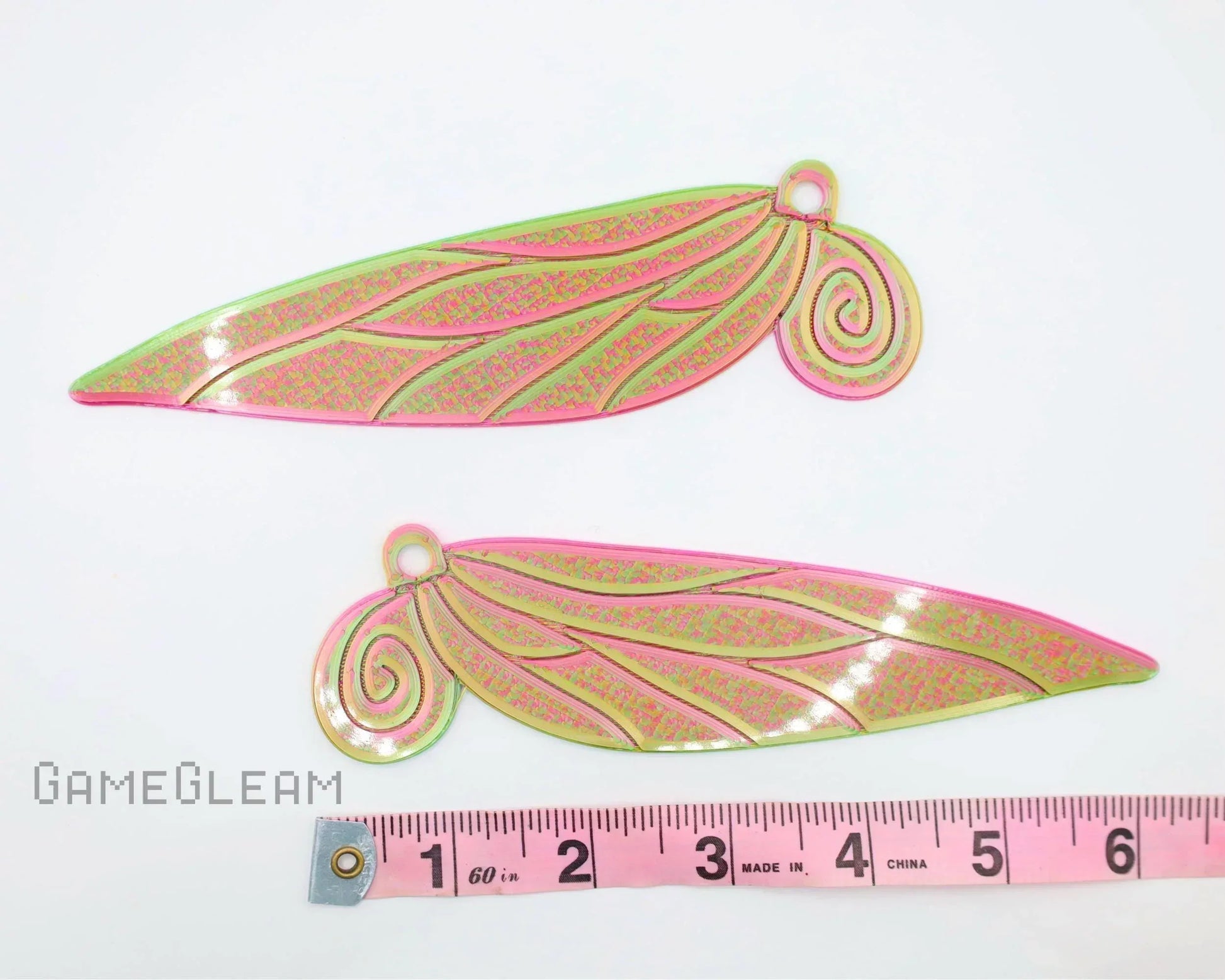Pair of pink and green holographic wing-shaped acrylic charms with swirl design, shown with measuring tape.