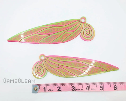 Pair of pink and green holographic wing-shaped acrylic charms with swirl design, shown with measuring tape.