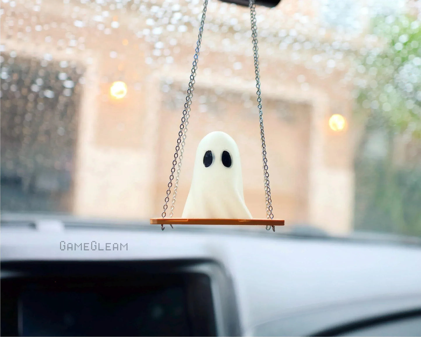 Handmade ghost car charm on swing with chain, hanging from rearview mirror, rainy window view