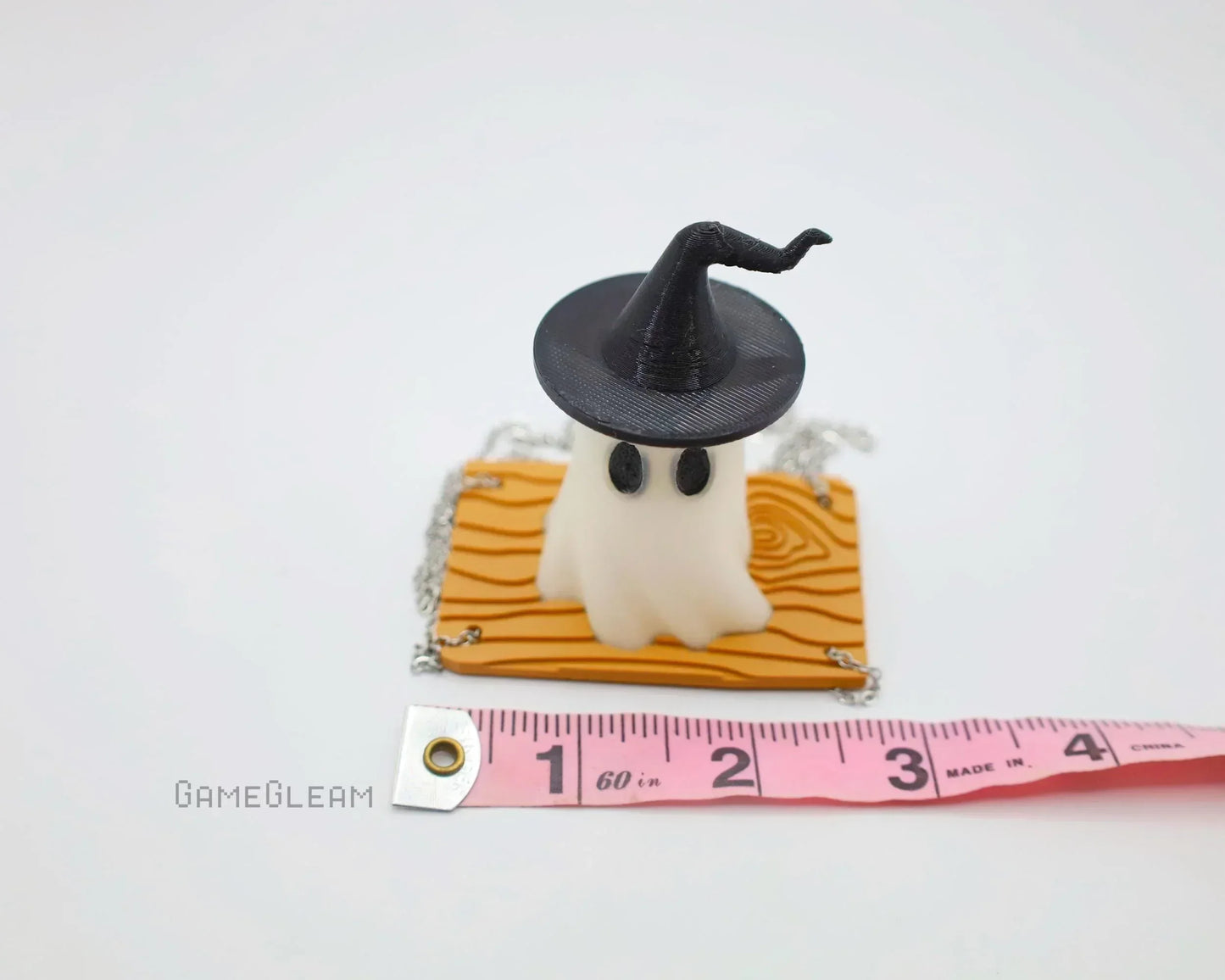 Handmade ghost figurine with witch hat on wood-texture base, shown next to tape measure