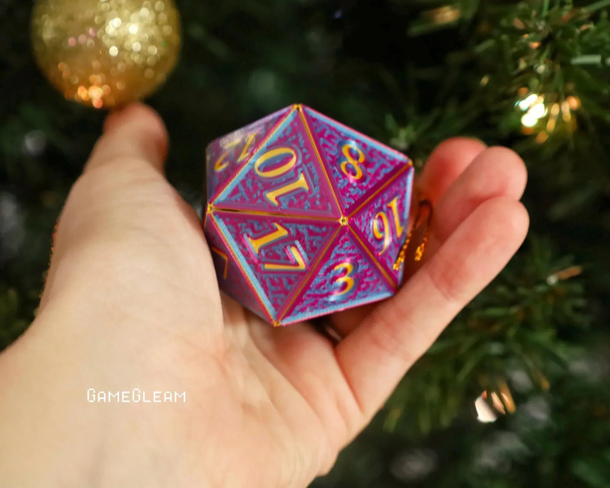 Hand holding holographic purple and gold twenty-sided dice ornament by a Christmas tree.