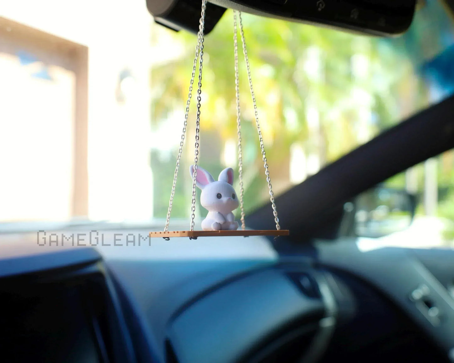 Cute white bunny car charm figurine on a swing, hanging from rearview mirror, in sunlight.