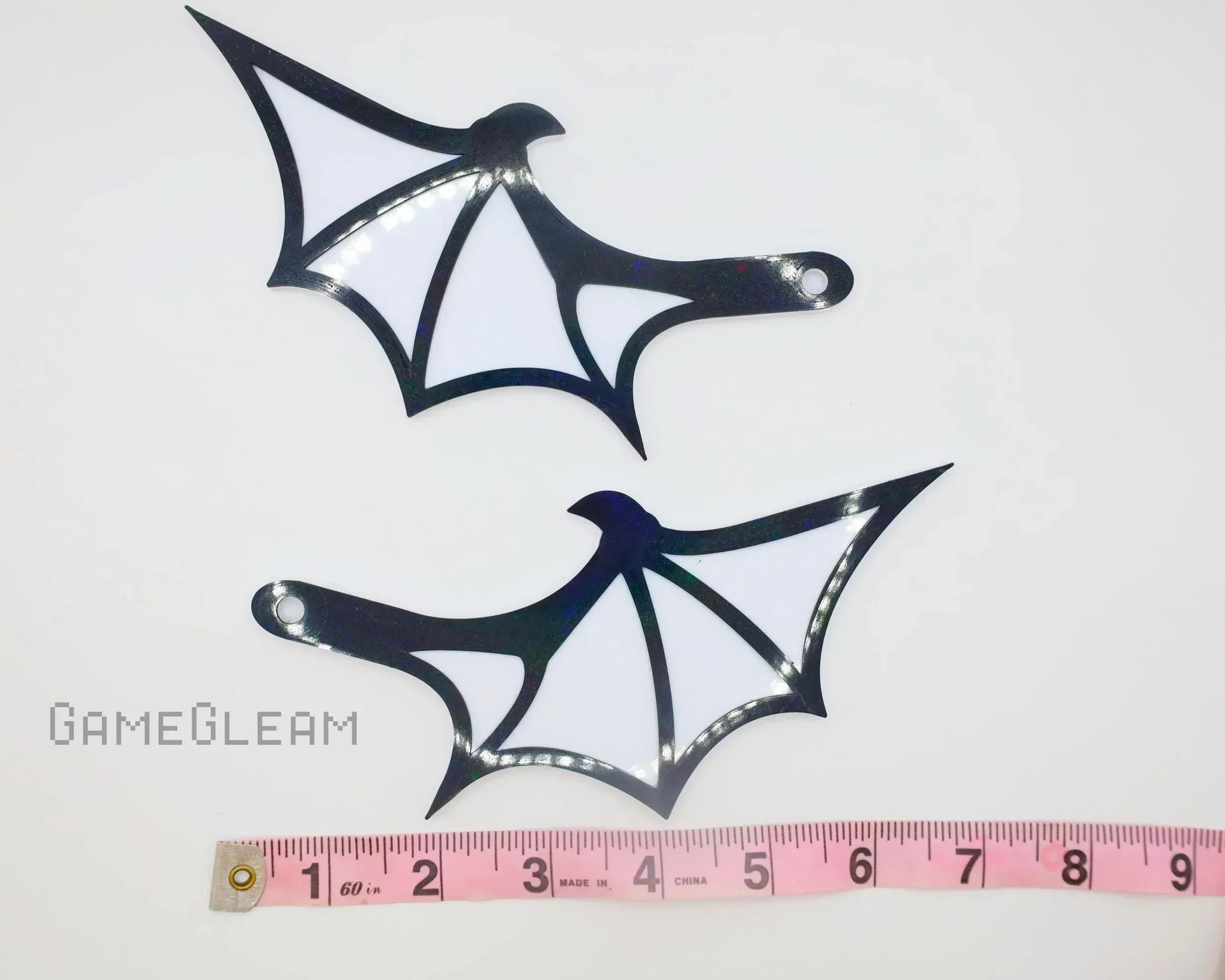 Two black and white bat wing-shaped acrylic charms with a measuring tape for scale