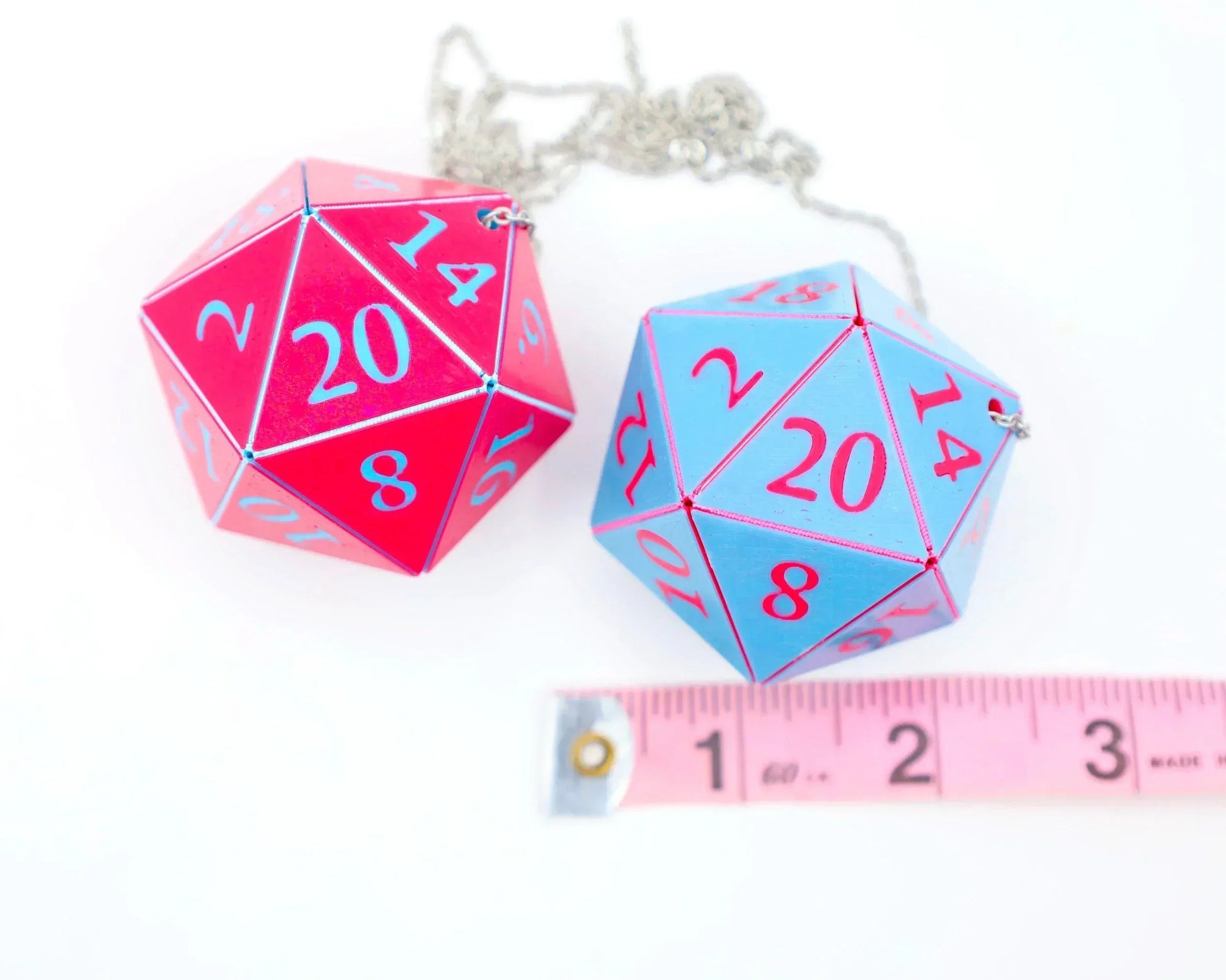 Large pink and blue 20-sided dice charms with chains beside a pink tape measure on white background