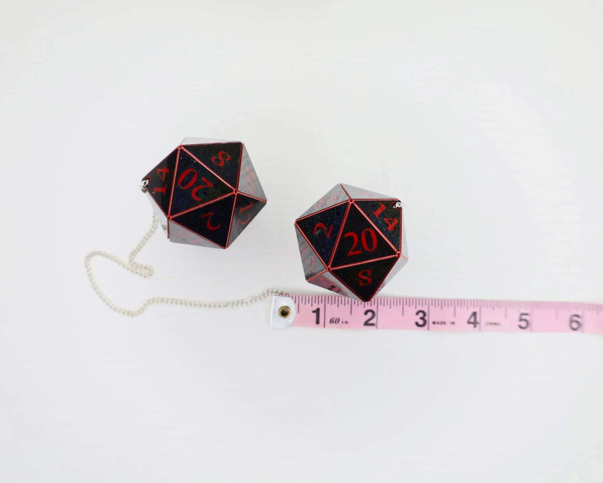 Two large black and red twenty-sided dice car charms with chain, next to a pink measuring tape