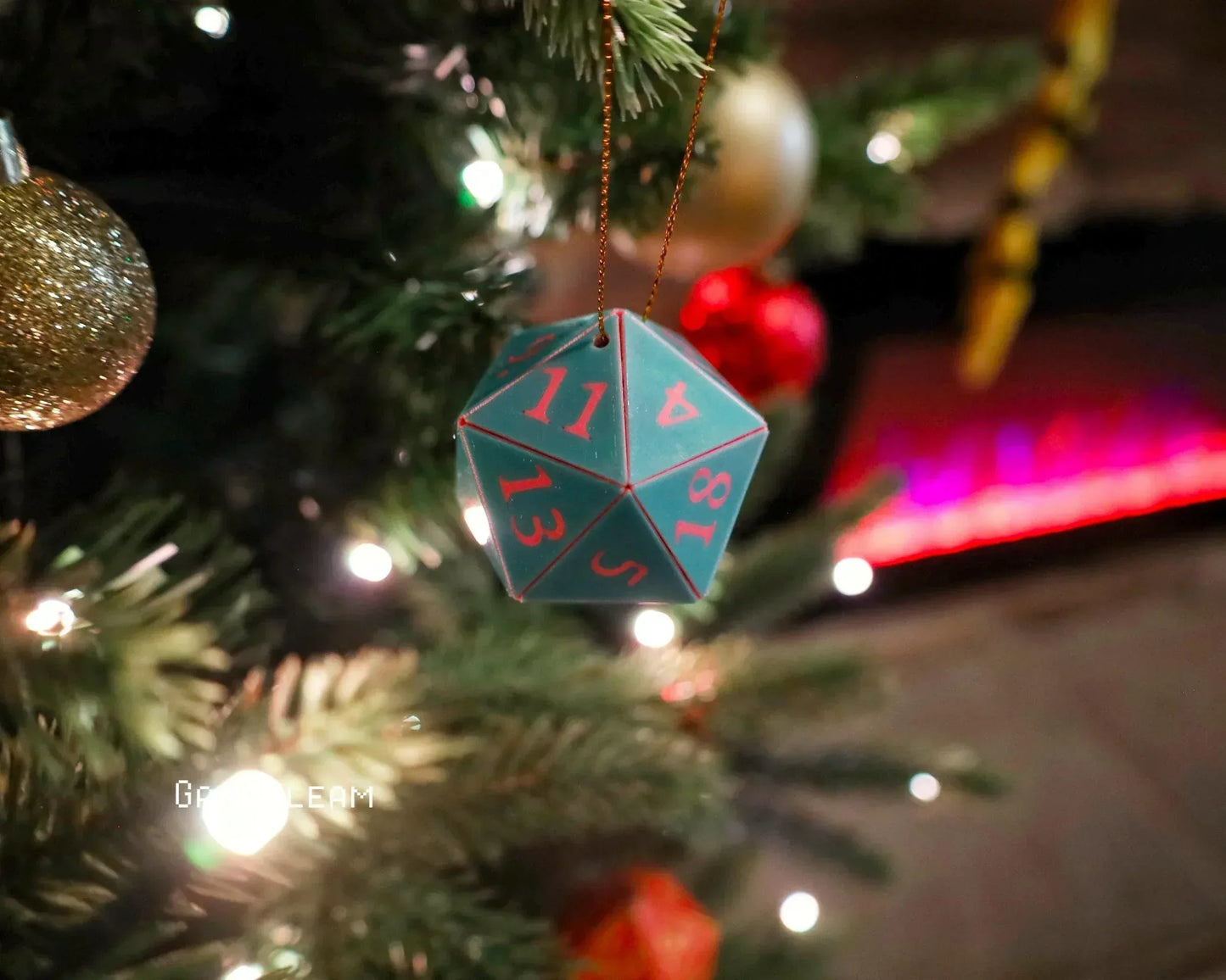 Teal twenty-sided dice ornament with red numbers hanging on a Christmas tree with festive lights