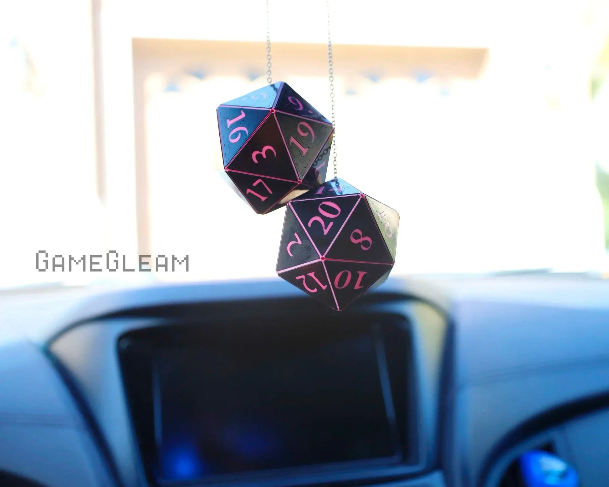Pink and black twenty-sided dice car charm hanging from rearview mirror in a car interior.
