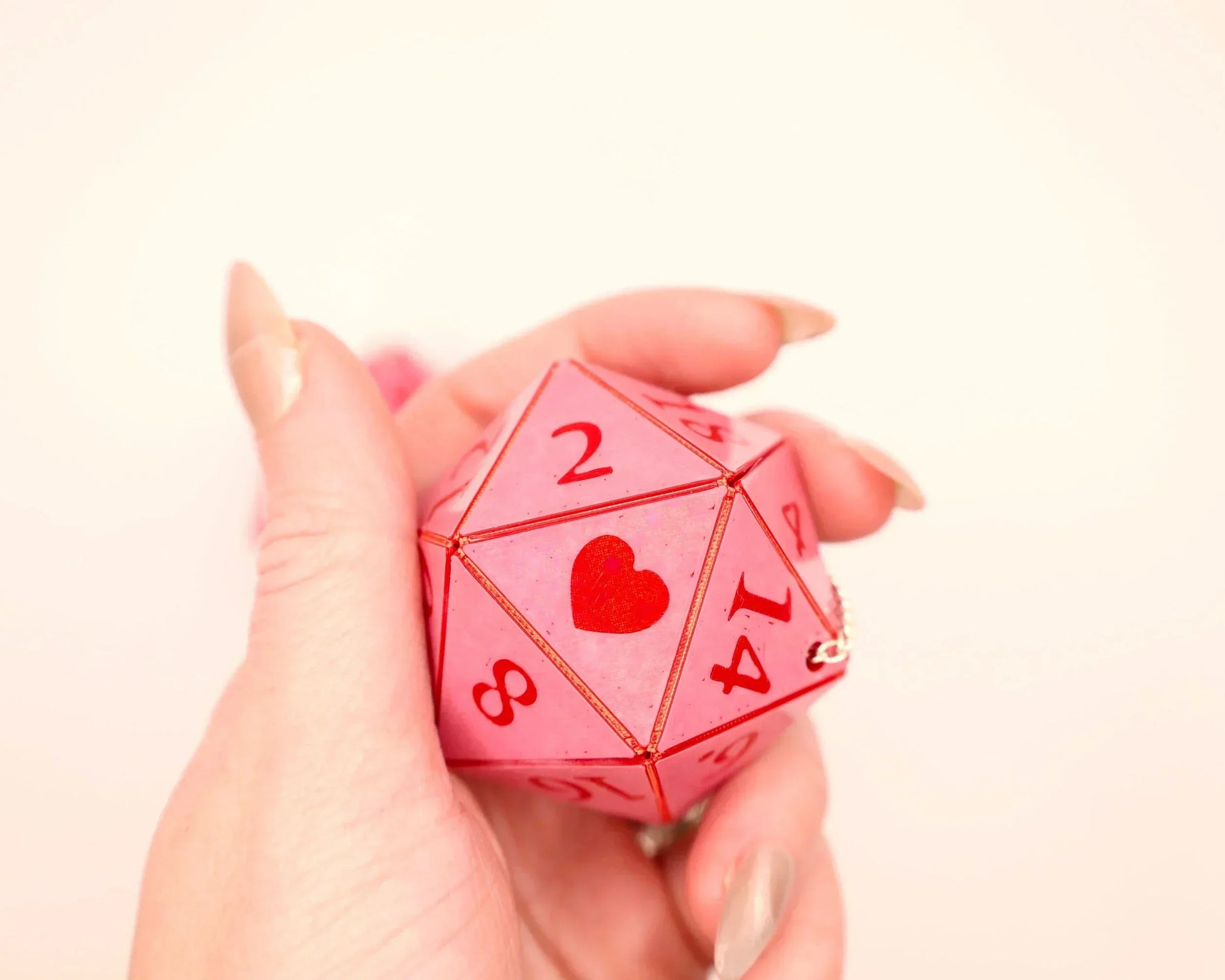 Hand holding a pink twenty-sided dice charm with red numbers and a heart symbol, gamer gift