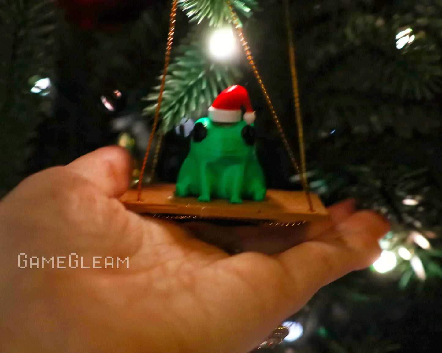 Hand holding a green frog Christmas ornament with Santa hat on a swing, near holiday tree.